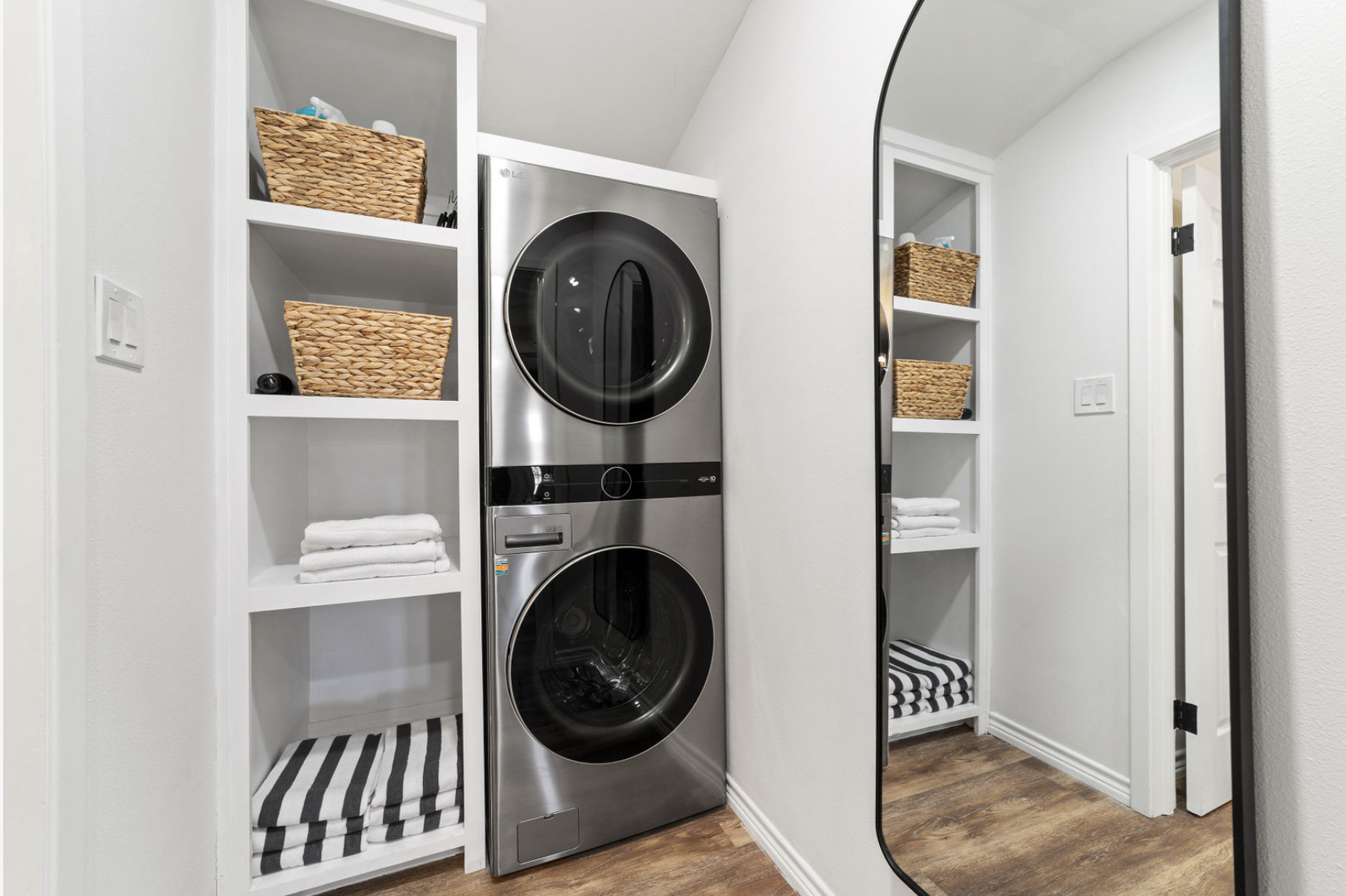A laundry room with white shelving on either side of a stainless steel washer and dryer, with woven baskets, folded towels, and a striped blanket, and a large mirror on the right wall.