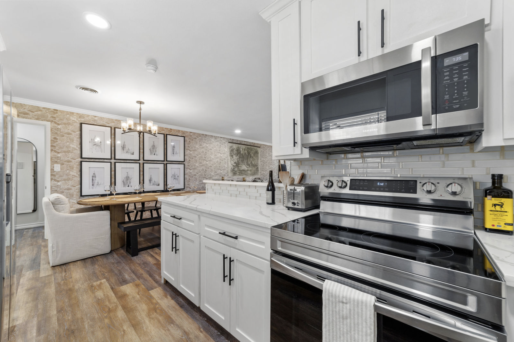 Modern kitchen with white cabinets, stainless steel appliances including an oven and microwave, a marble countertop, and a dining area with a wooden table and mismatched chairs. The wall features framed artwork, and the space has a rustic brick accen