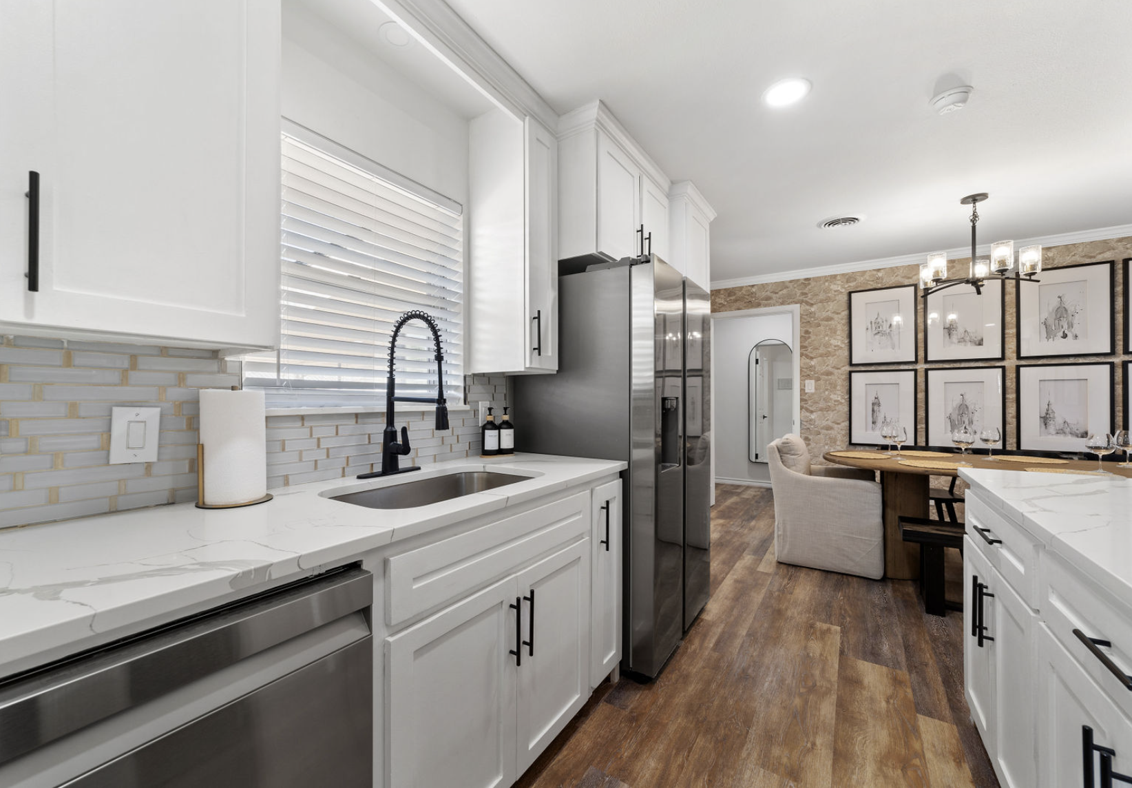 Modern kitchen with white cabinets, black hardware, stainless steel appliances, marble countertops, a black farmhouse sink with a high-arc black faucet, window with blinds, and a dining area with a wooden table, beige chairs, and framed artwork on a 