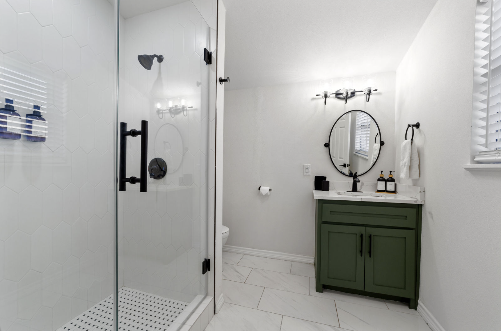 A modern bathroom with a glass shower enclosure, black fixtures, white hexagon tile walls, and a white vanity with a green cabinet. There is a round mirror above the sink, with black fixtures and accessories, including soap dispensers, a towel, and a