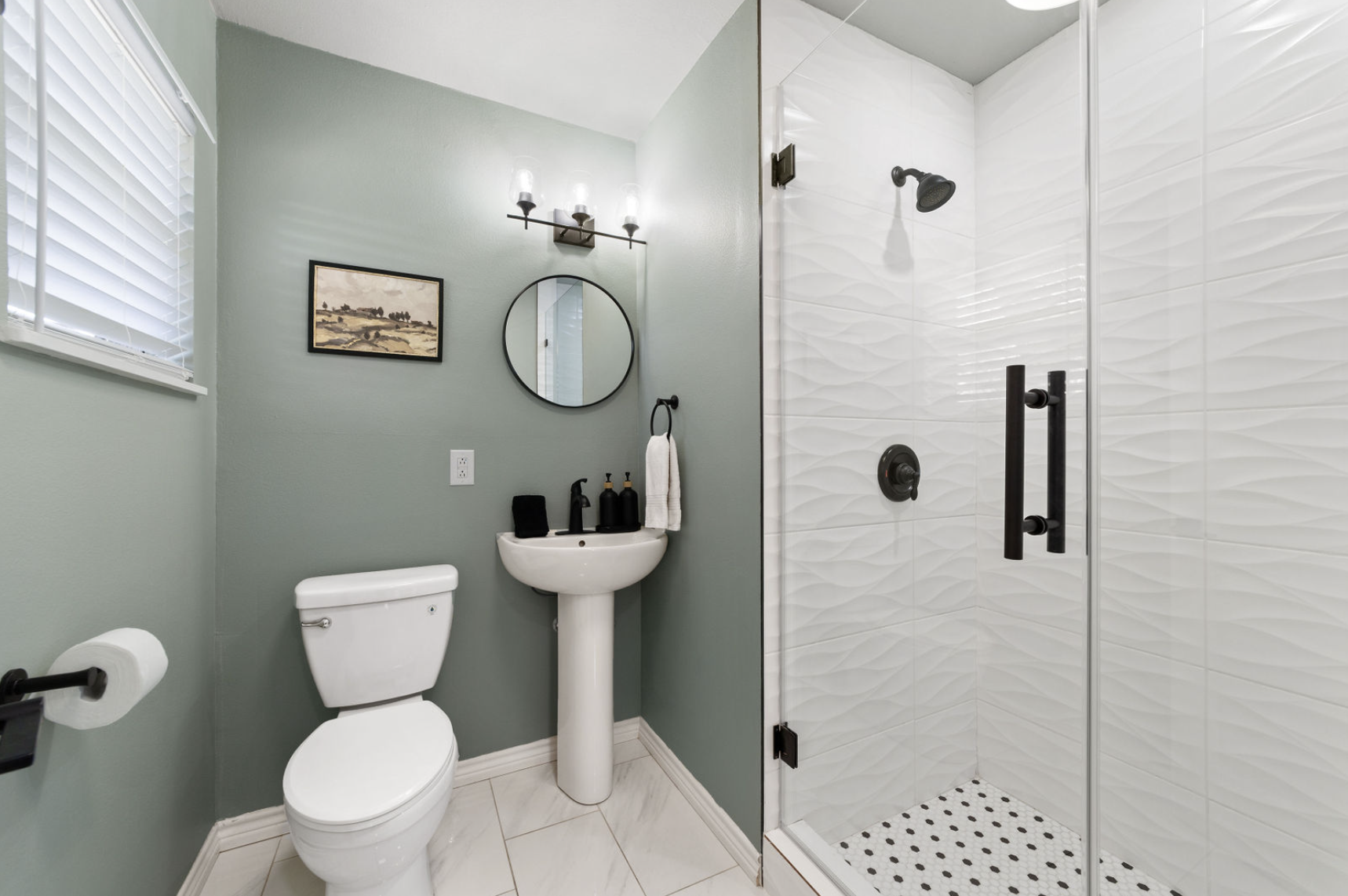A small bathroom with a toilet, a pedestal sink with soap dispensers, a round mirror, a wall-mounted light fixture, a picture on the wall, a towel ring with a white towel, and a glass-enclosed shower with black fixtures and textured white tiles.