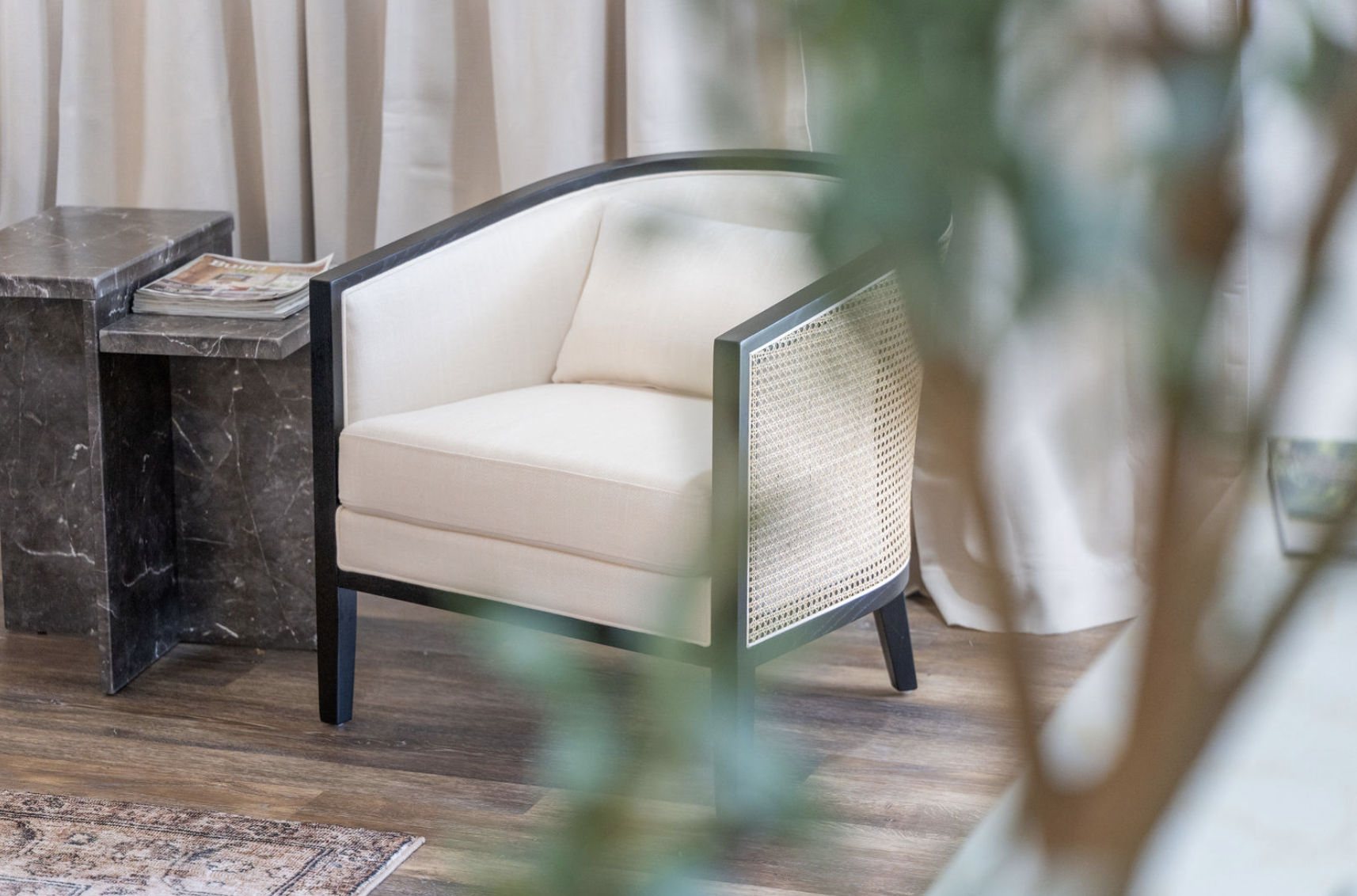 A cream-colored armchair with a black frame and woven backrest, placed next to a black marble side table with magazines. The room has wooden flooring and cream-colored curtains in the background, with a soft plant partially visible in the foreground.
