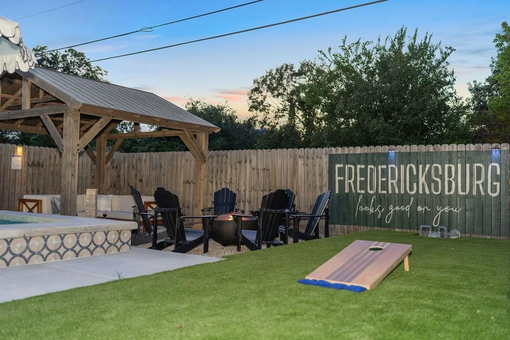Backyard with a wooden pavilion, Adirondack chairs around a fire pit, a cornhole game on the grass, and a fence with a sign that says "FREDERICKSBURG looks good on you" during early evening.