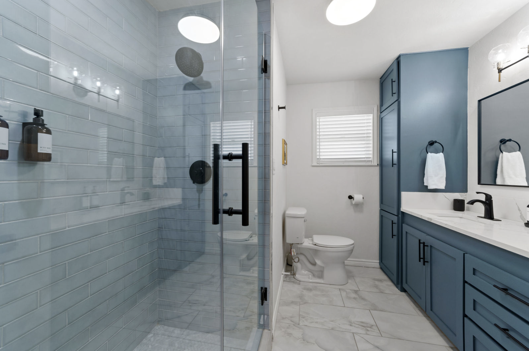 A modern bathroom with a glass-enclosed shower, a white toilet, blue cabinetry with black handles, a marble countertop, a black faucet, a rectangular mirror, and white walls with window blinds.