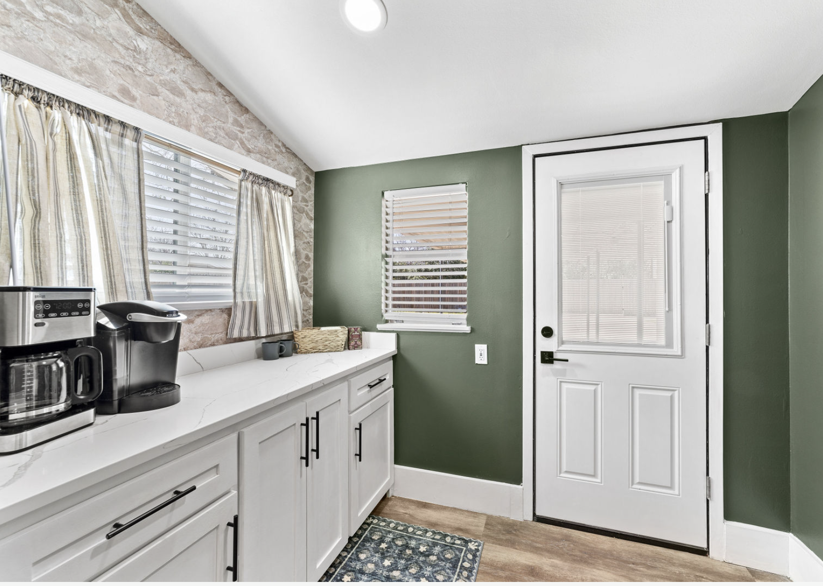 Kitchen with white cabinets, green wall, window with curtains, coffee maker, toaster, small rug, white door with window, wood flooring.