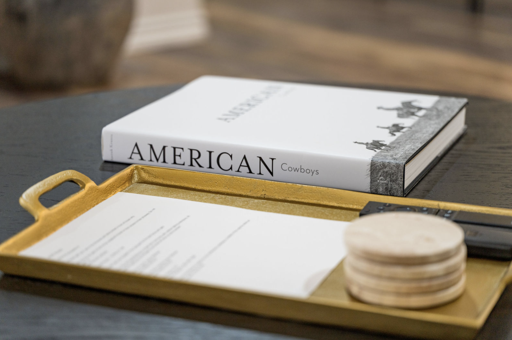 A hardcover book titled 'American Cowboys' resting on a dark table, with part of a yellow serving tray and a remote control visible nearby.