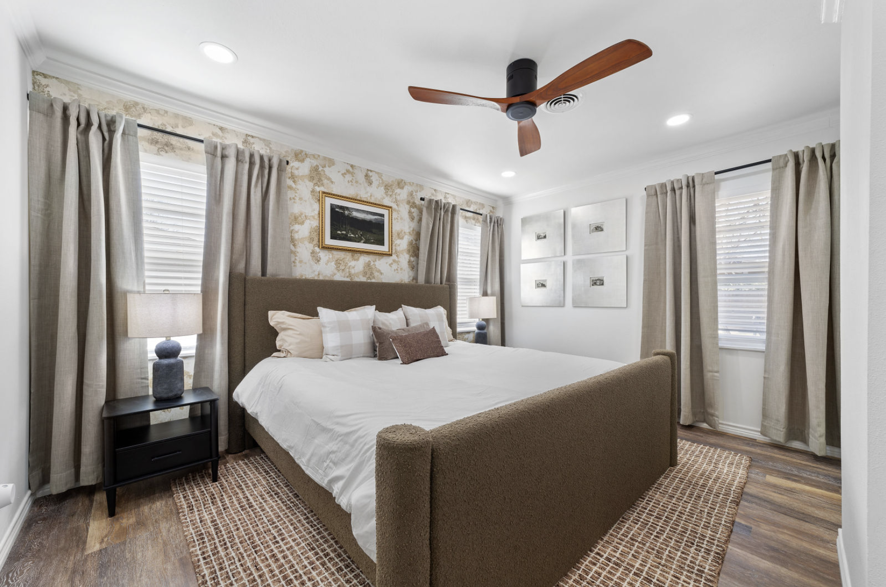 A neatly made bedroom with a bed that has a brown upholstered headboard and a variety of pillows. Flanking the bed are two black nightstands, each with a blue ceramic table lamp. The room features a white ceiling with recessed lighting, a ceiling fan with wooden blades, and a wall decorated with framed artwork and photographs. Four windows with white blinds and beige curtains allow natural light into the room. The floor has wood flooring with a patterned area rug.