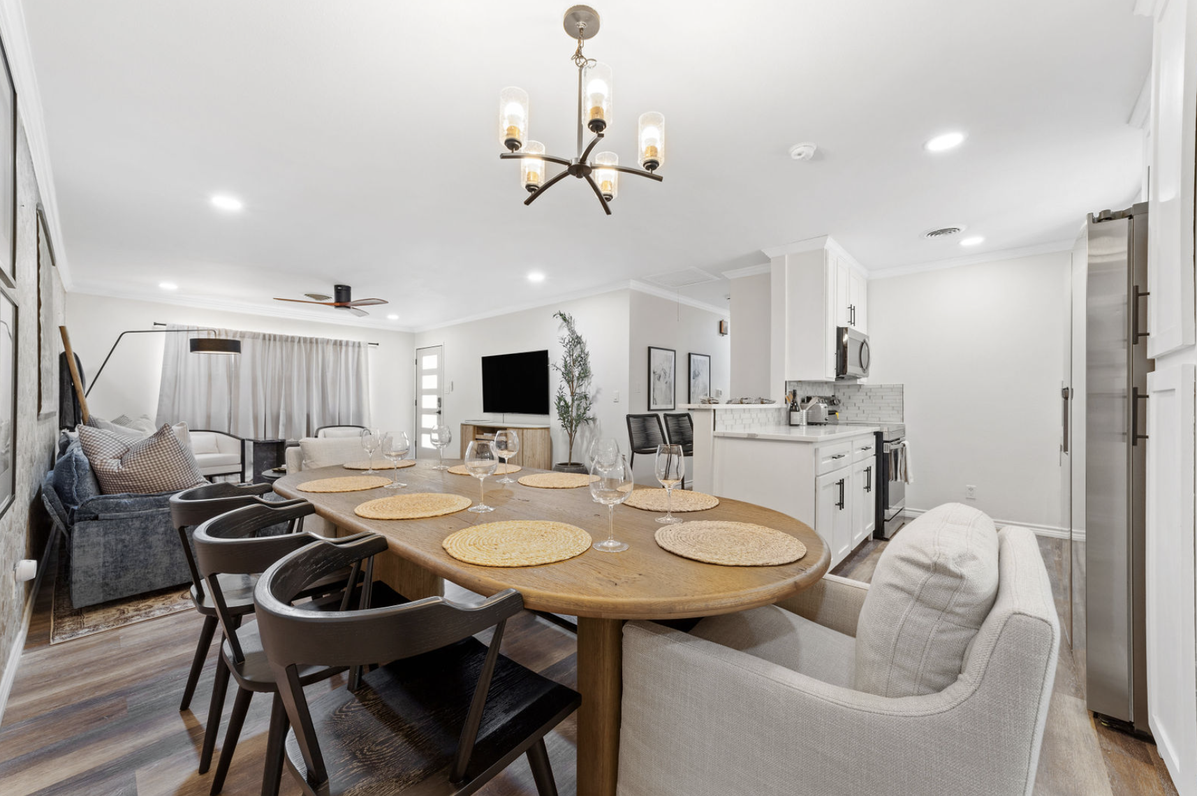Open-concept living and dining area with a kitchen, dining table set with placemats and wine glasses, a sofa with pillows, a wall-mounted TV, and modern decor.