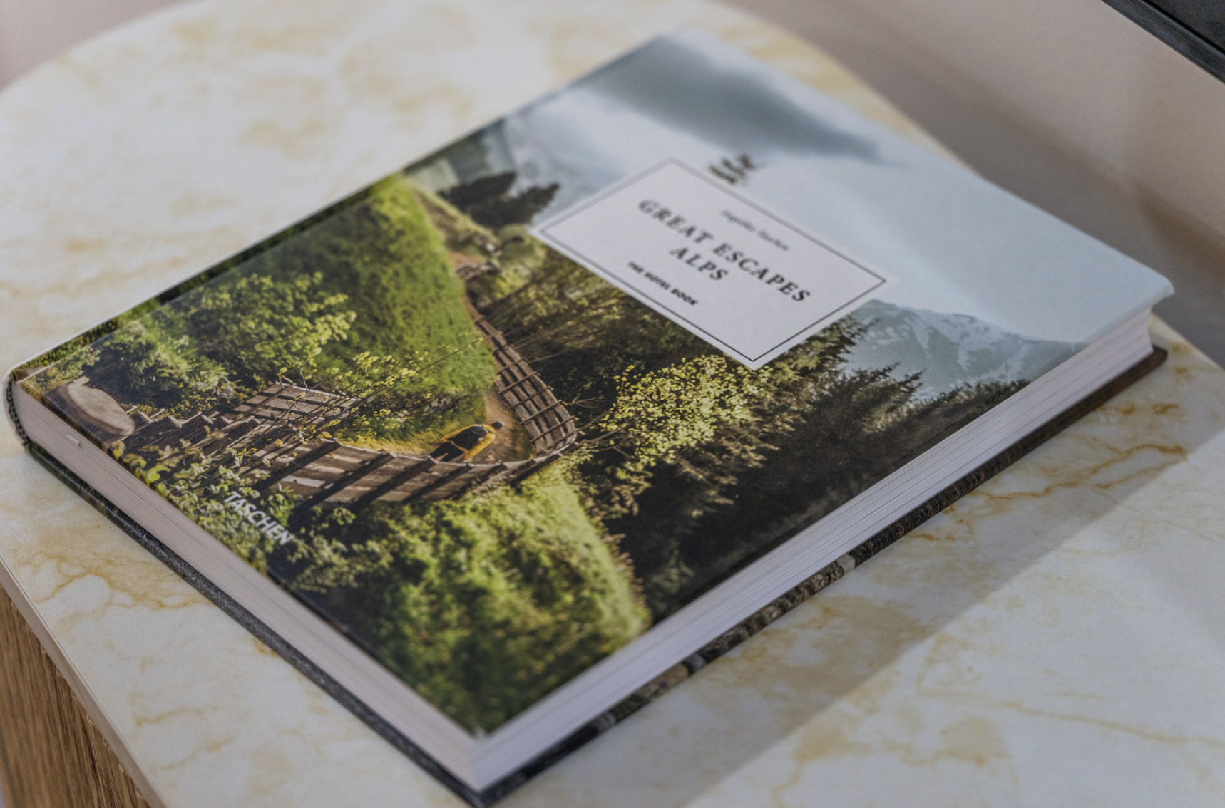 A travel guidebook titled "Great Escapes: Alps" lying on a marble table. The cover features a scenic photograph of lush green trees, a wooden fence, and mountain peaks in the background.