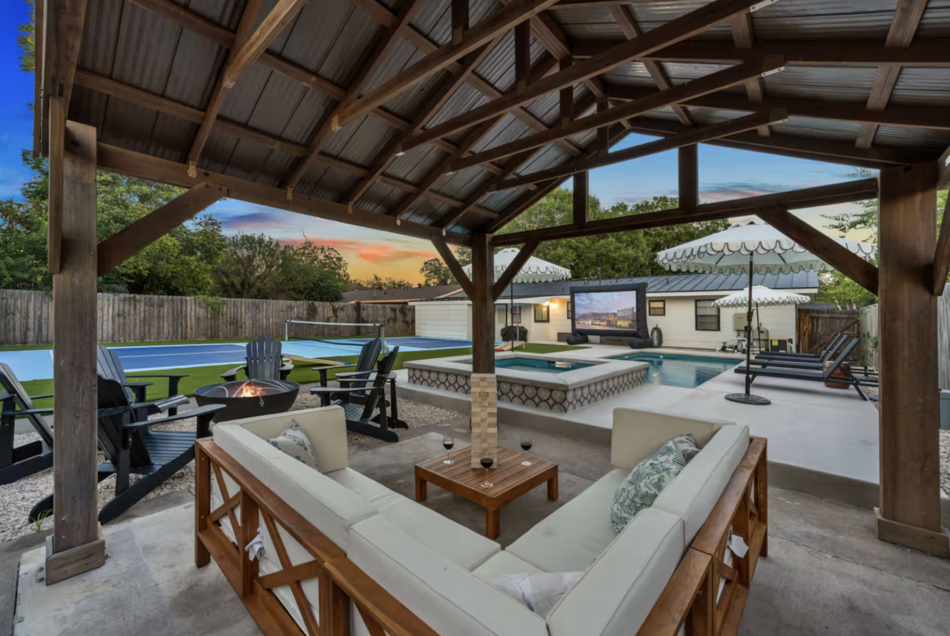 Backyard patio area with a covered wooden pergola, seating area, hot tub, swimming pool, umbrellas, and a large screen television at sunset.