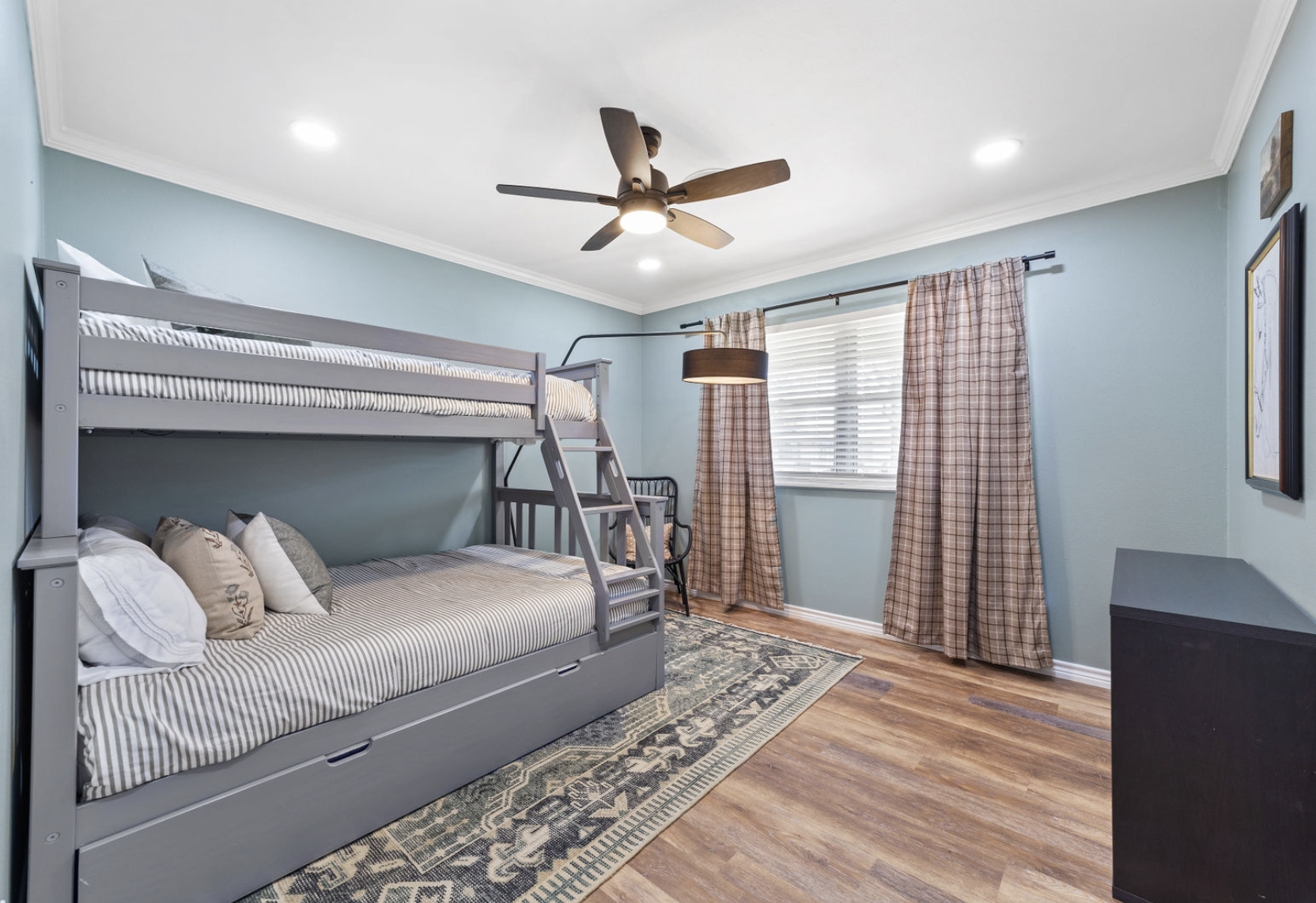 A room with light blue walls, a bed with gray bunk bed frame and striped bedding, multiple pillows, a window with white blinds and plaid curtains, a ceiling fan with wooden blades, a black chair, a beige patterned rug, and a black dresser.