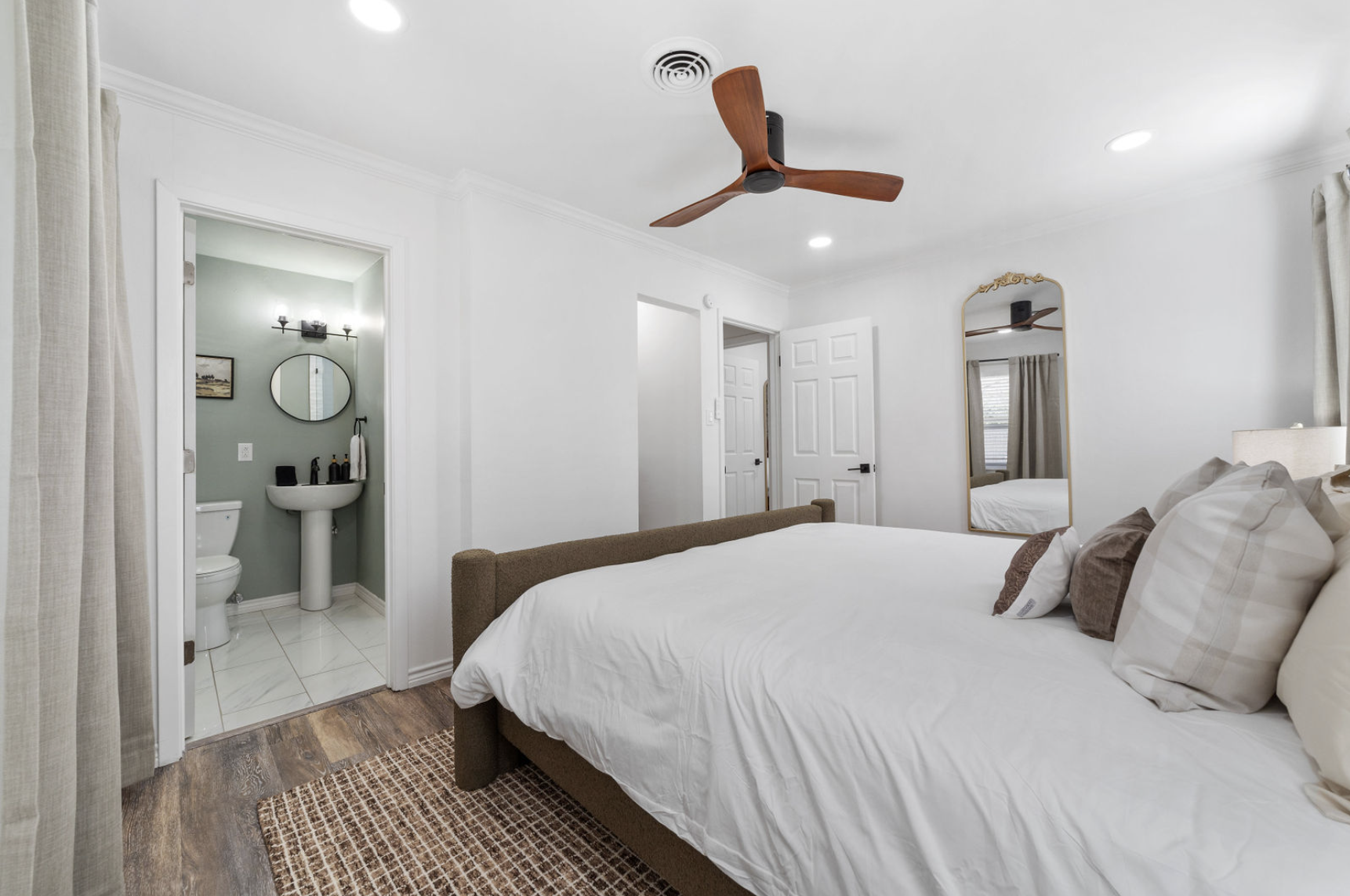 A bedroom with a large bed with white linens and multiple pillows, a mirror on the wall, a ceiling fan, and an adjoining bathroom with a pedestal sink, mirror, and toilet.