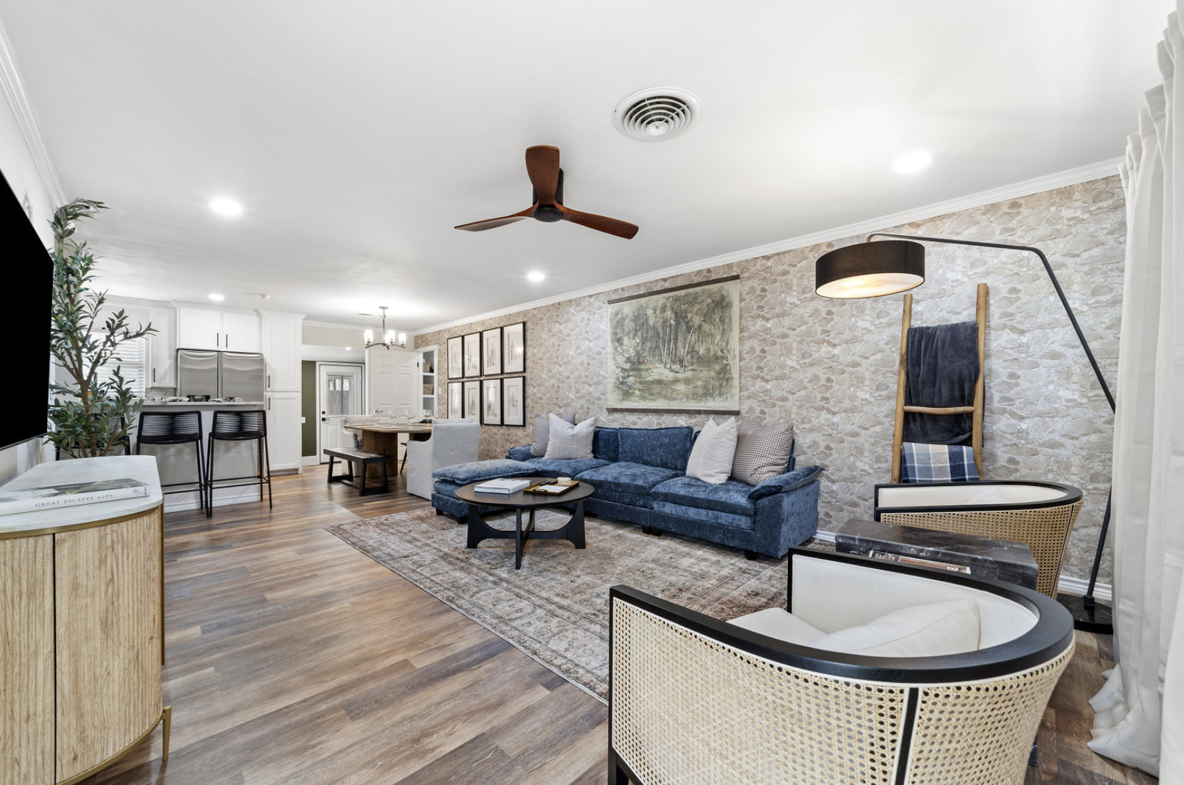 Open-concept living room with a blue velvet sofa, various armchairs, a coffee table, wall art, a stone accent wall, and a view into the kitchen area.