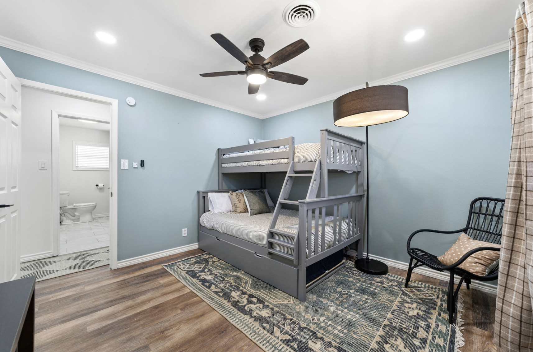 A bedroom with a gray bunk bed, light blue walls, a black chair with a beige pillow, a rug, a standing lamp with a brown shade, ceiling fan with dark blades, and an adjoining bathroom with a toilet and window.