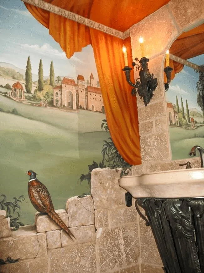 Decorative interior wall with painted landscape scene, stone wall accents, orange curtains, black wrought iron wall sconce with candle, and a ceramic bird figurine near the window.