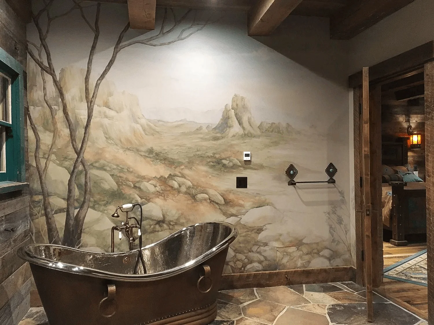 A rustic bathroom with a large, vintage, copper bathtub with clawfoot legs, situated in front of a mural depicting a desert landscape with mountains and rocky terrain.