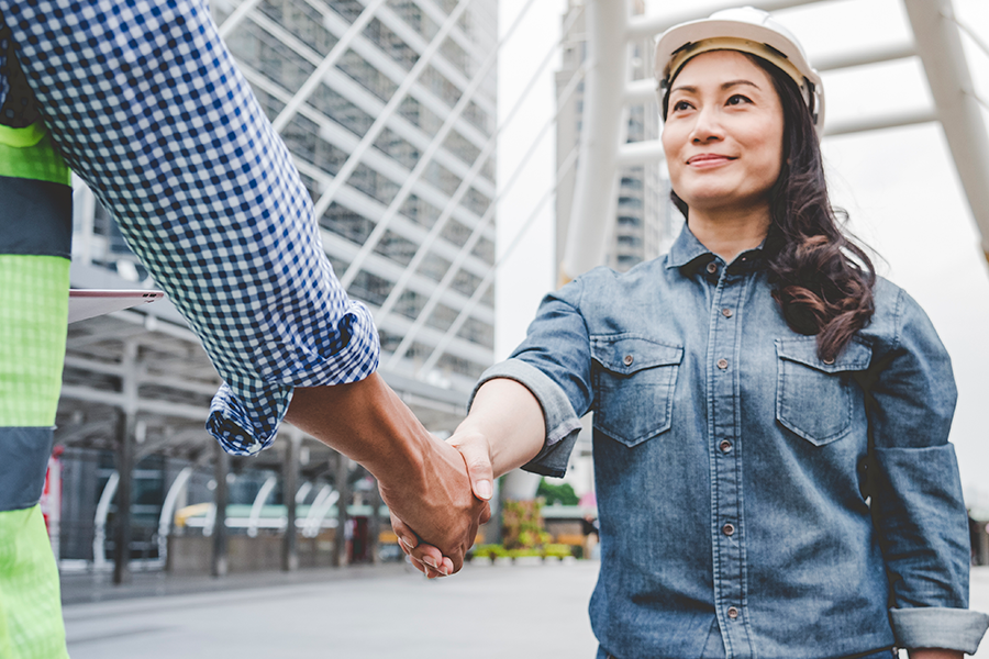 A woman wearing a safety helmet shaking hands with a person in a high-visibility vest in an urban construction or development area.