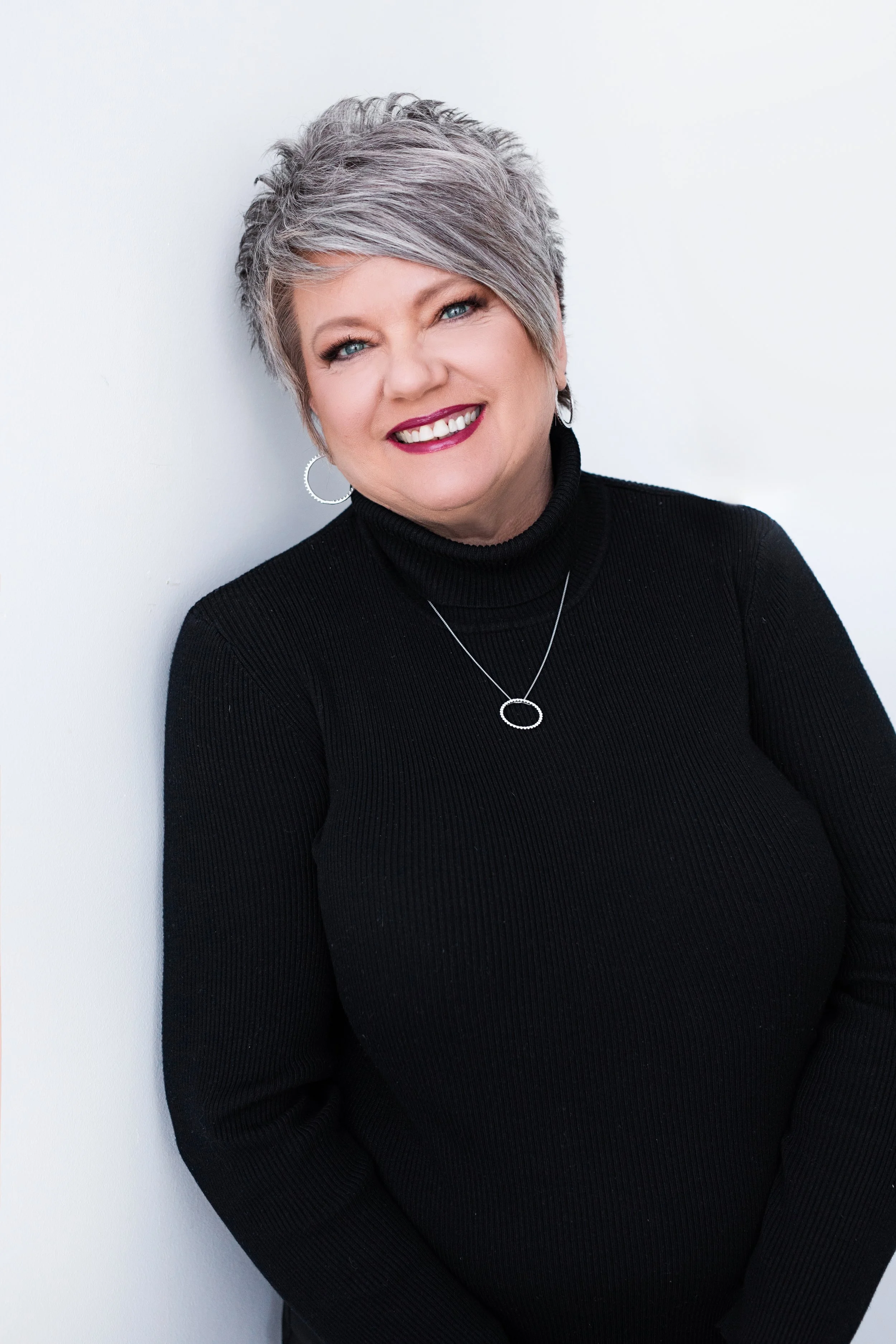 A smiling woman with short gray hair wearing a black turtleneck sweater, silver hoop earrings, and a silver necklace with a circular pendant, standing against a white background.