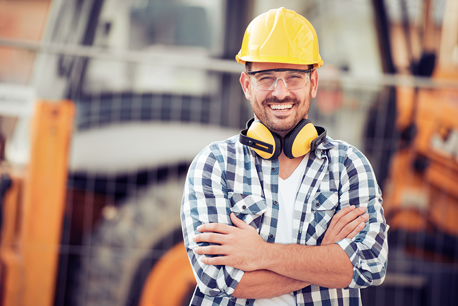 A smiling man wearing a yellow safety helmet, clear safety glasses, and yellow earmuffs around his neck, standing on a construction site with orange equipment and a building in the background.