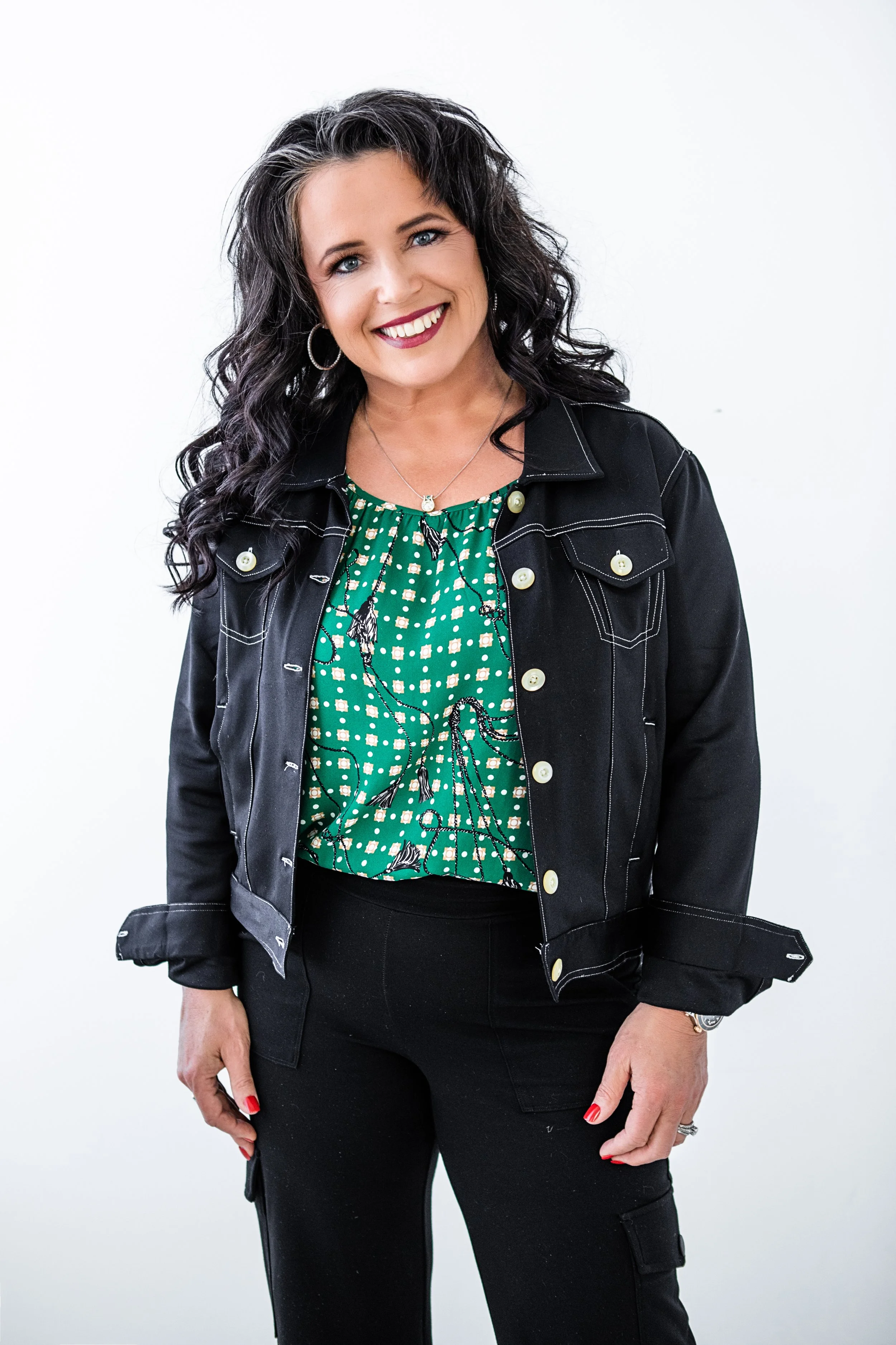A woman with shoulder-length curly black hair, blue eyes, and red lipstick, smiling and wearing a black denim jacket over a green top with a white and black pattern. She has hoop earrings, a necklace, and a watch, and is standing against a plain white background.