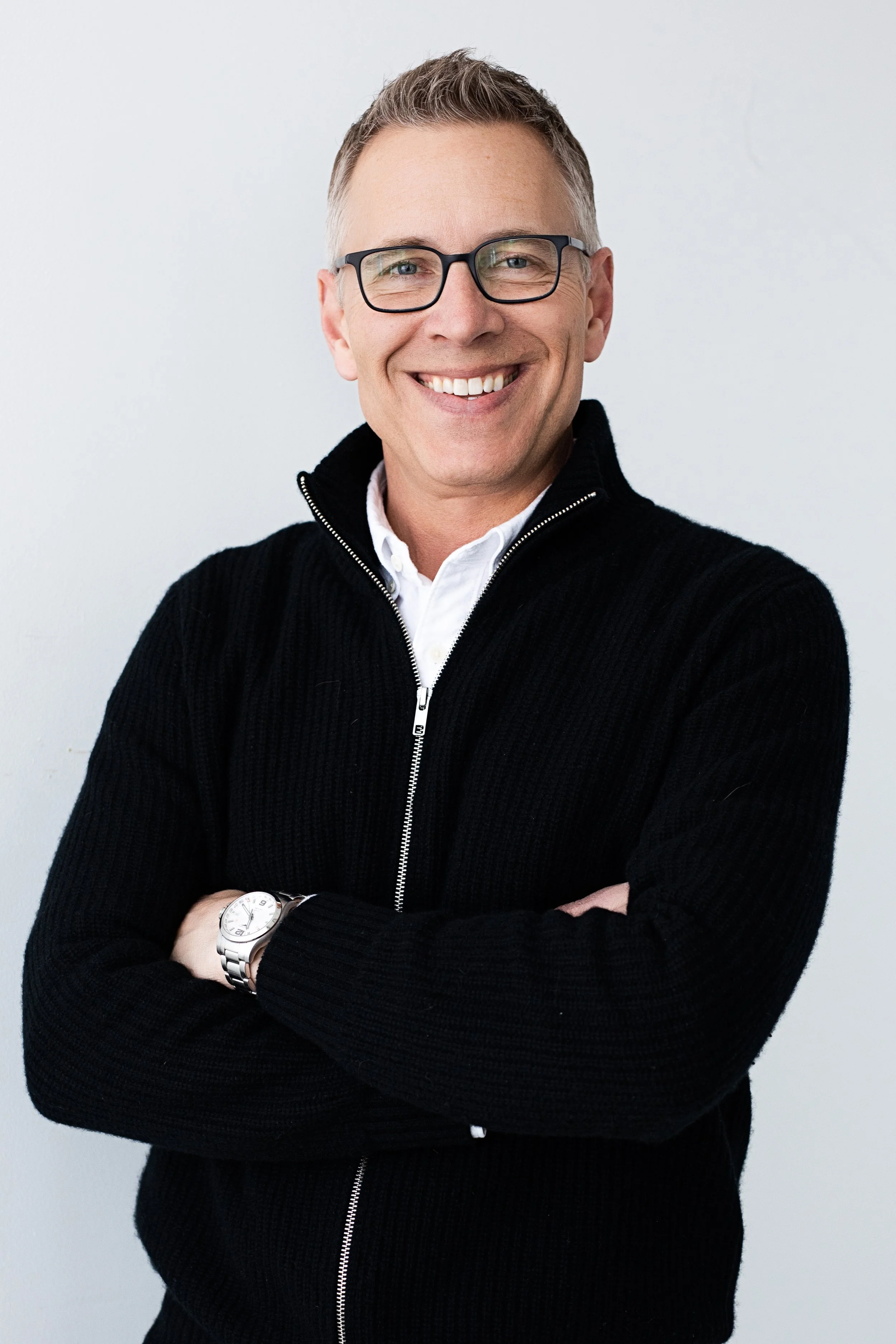 A man with short hair, glasses, and a smile wearing a black zip-up sweater over a white shirt, standing against a plain white background.