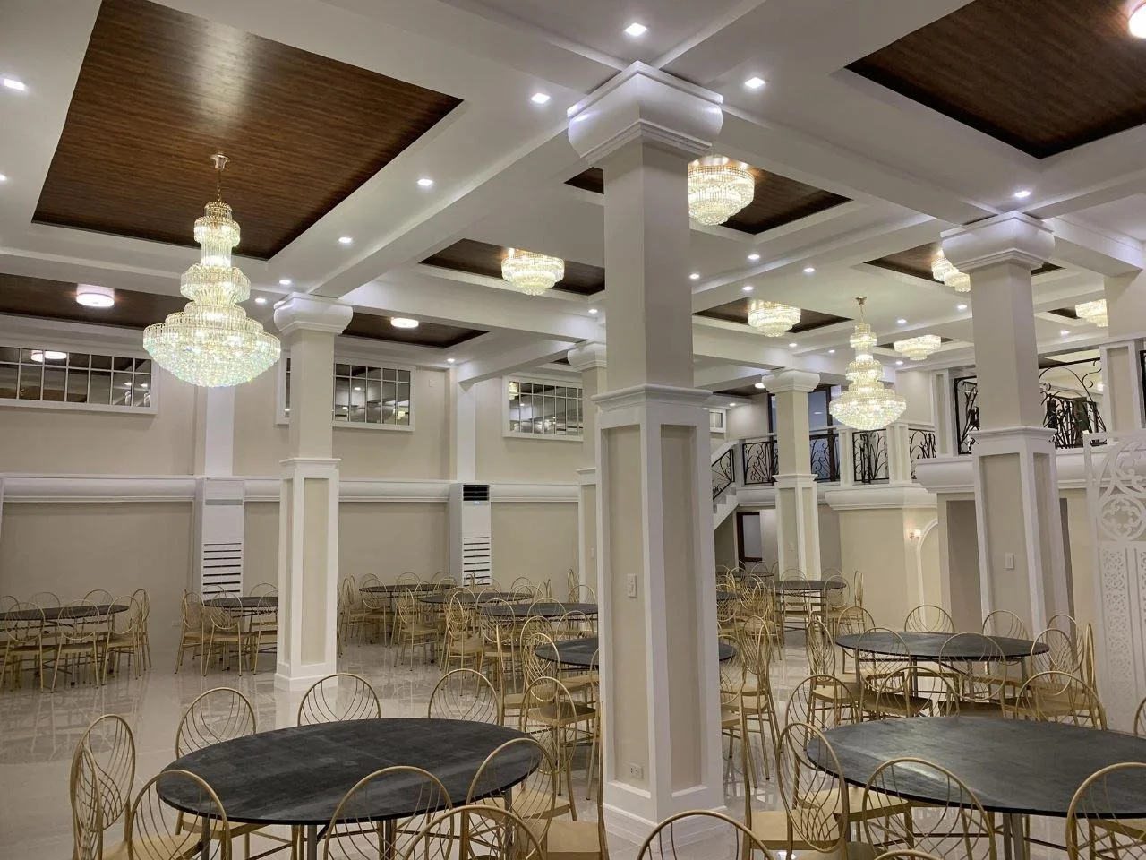 Elegant banquet hall with chandeliers, white walls, dark wood ceiling accents, gold chairs, black tables, and a staircase with wrought iron railings.