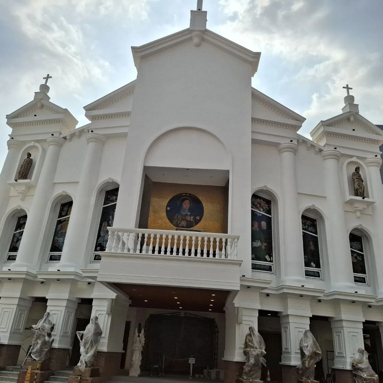 Front view of a white church with statues in niches, stained glass windows, a circular painting in the center, and guards covered with plastic outside.