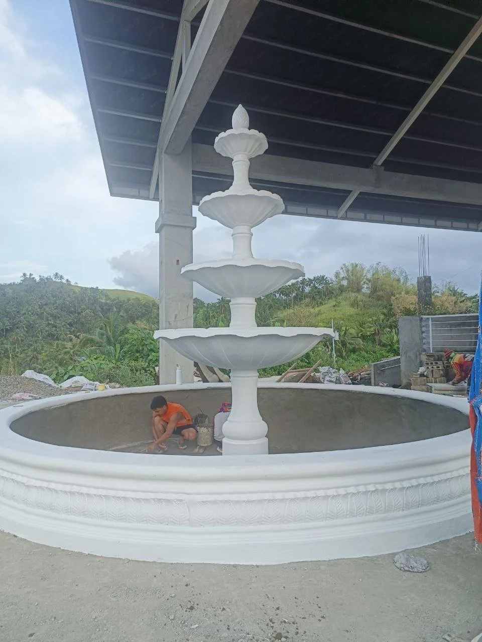 A four-tier white fountain under construction in an open area with a green hillside and cloudy sky in the background, and workers nearby.