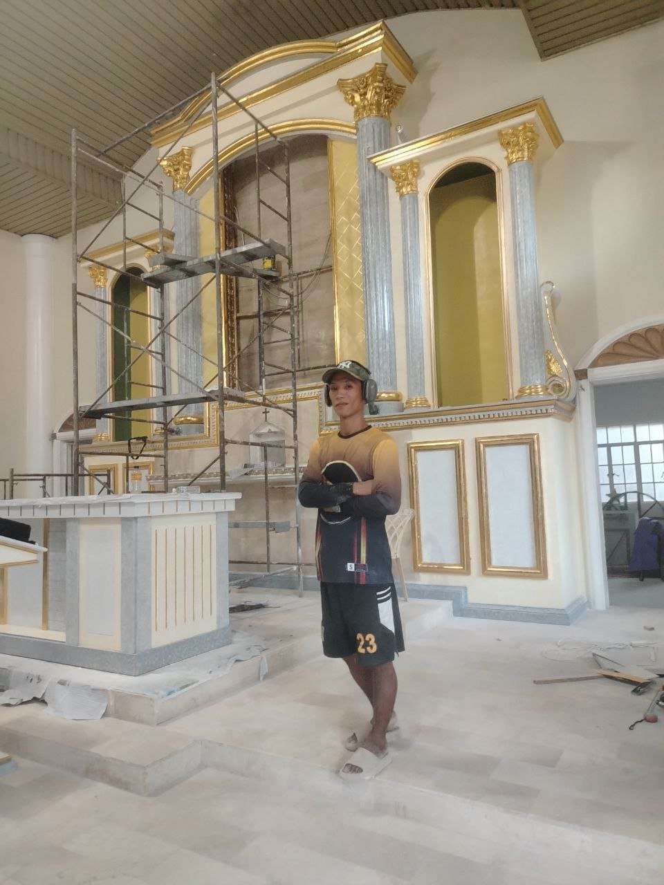 A man standing inside a church under renovation, wearing headphones, a long-sleeved shirt, shorts with the number 23, and gloves, with scaffolding and ornate gold and gray columns and panels in the background.