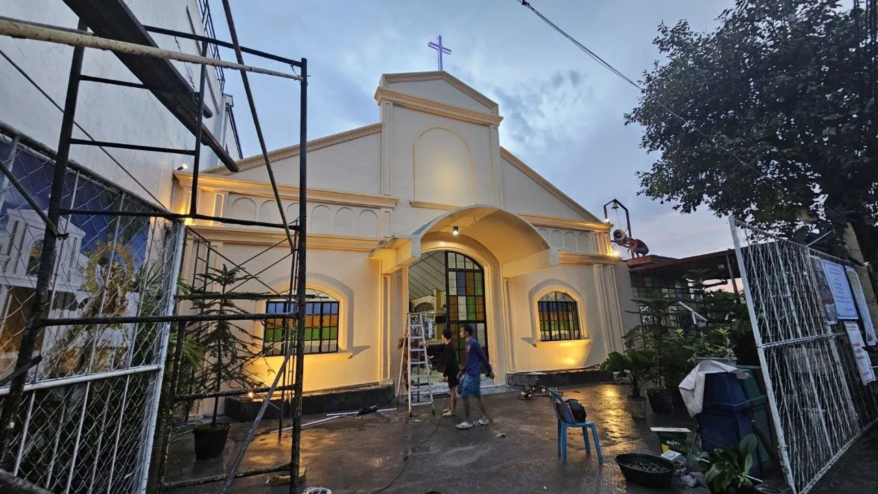 Front view of a church building with a lit cross on top, surrounded by construction equipment and scaffolding, during sunset.