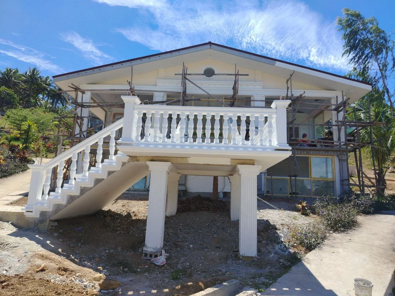 A house under construction with white pillars, a staircase, and a balcony, surrounded by scaffolding and greenery under a blue sky.