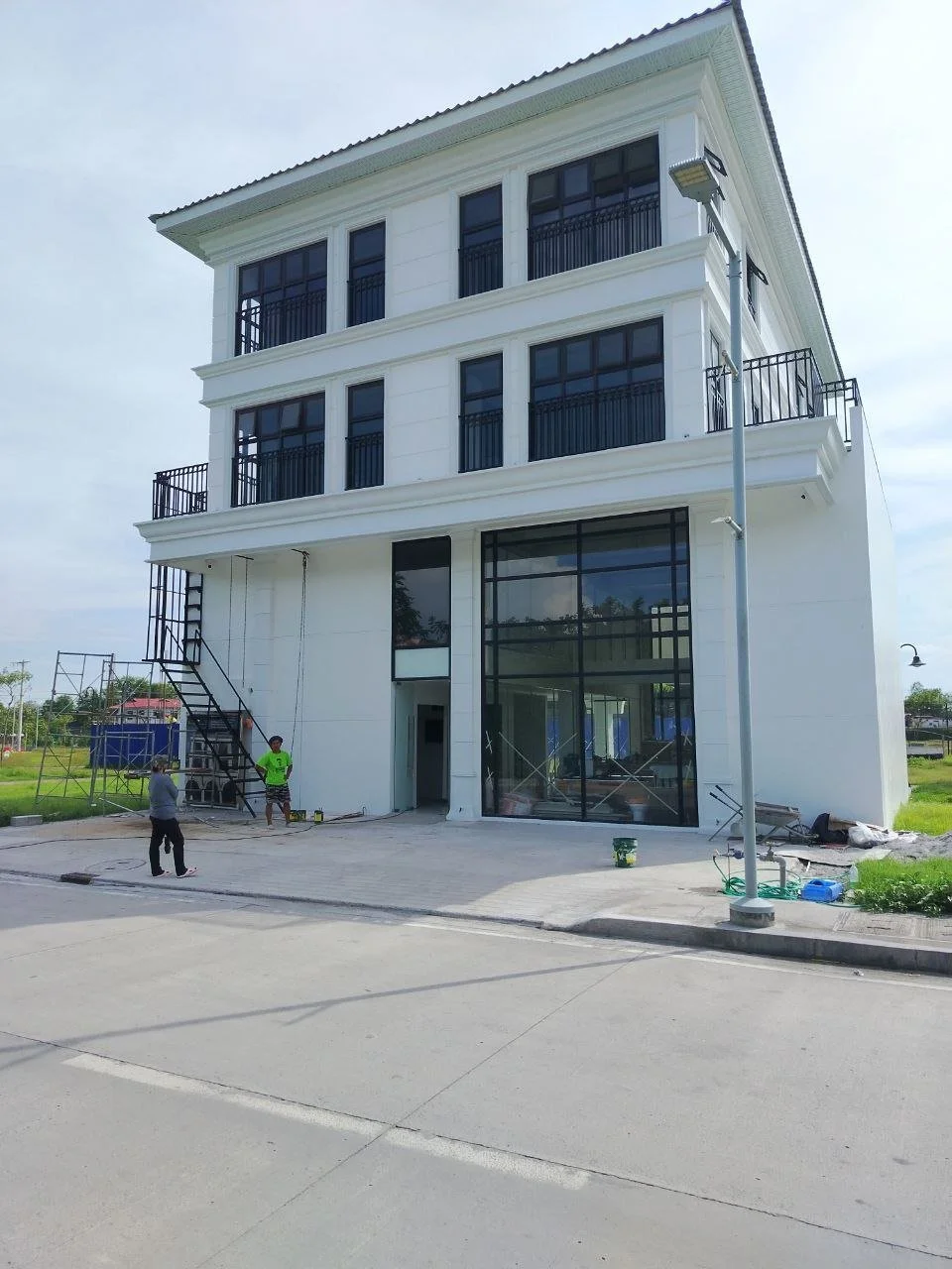 New white multi-story building under construction with workers on site, large glass windows, and balconies with black railings, next to a street with a streetlamp.