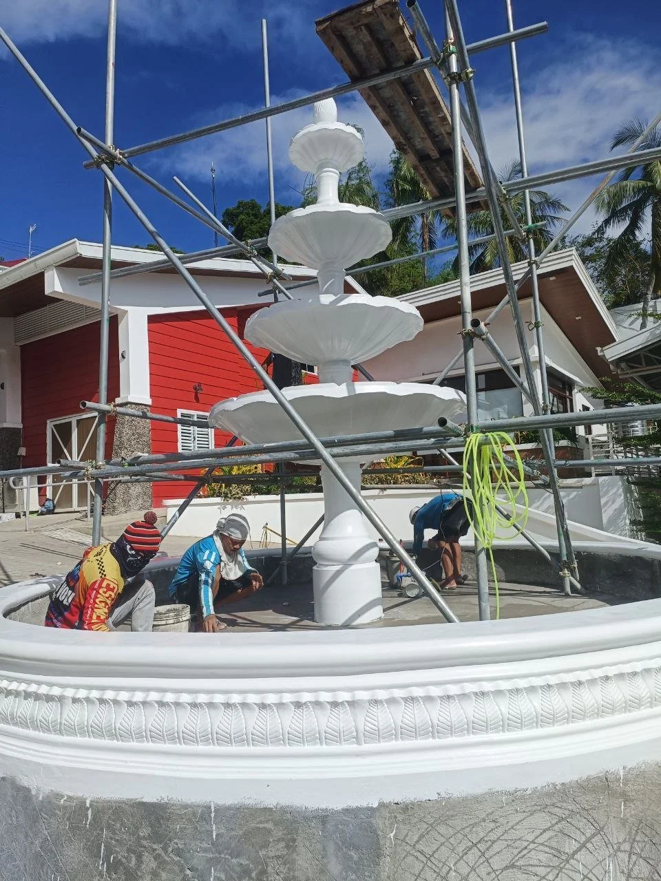 Workers installing or repairing a white, decorative fountain with scaffolding around it in a residential area with palm trees and a red house.