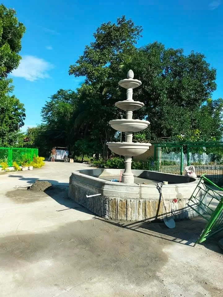 A multi-tiered white fountain in a garden area with trees and a blue sky. There are some construction materials and a green bicycle nearby.