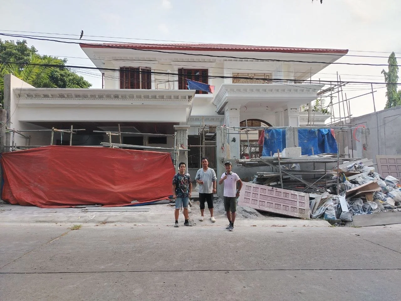 Three men standing in front of a large house under construction, with scaffolding, building materials, and debris around.