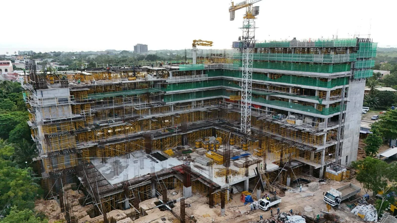 A multi-story building under construction with scaffolding, yellow exterior support structures, green safety barriers, a large construction crane, and construction vehicles on site.