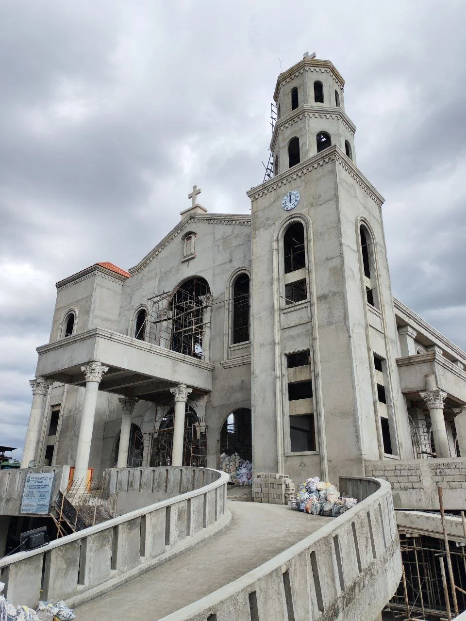 A church under construction with a gray stone facade, scaffolding on the windows, and piles of construction materials outside.
