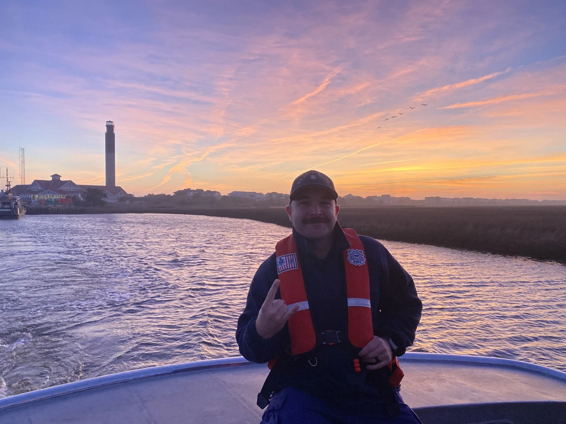 Person with mustache wearing a life jacket and cap on a boat during sunset, with a lighthouse, houses, and flying birds in the background.