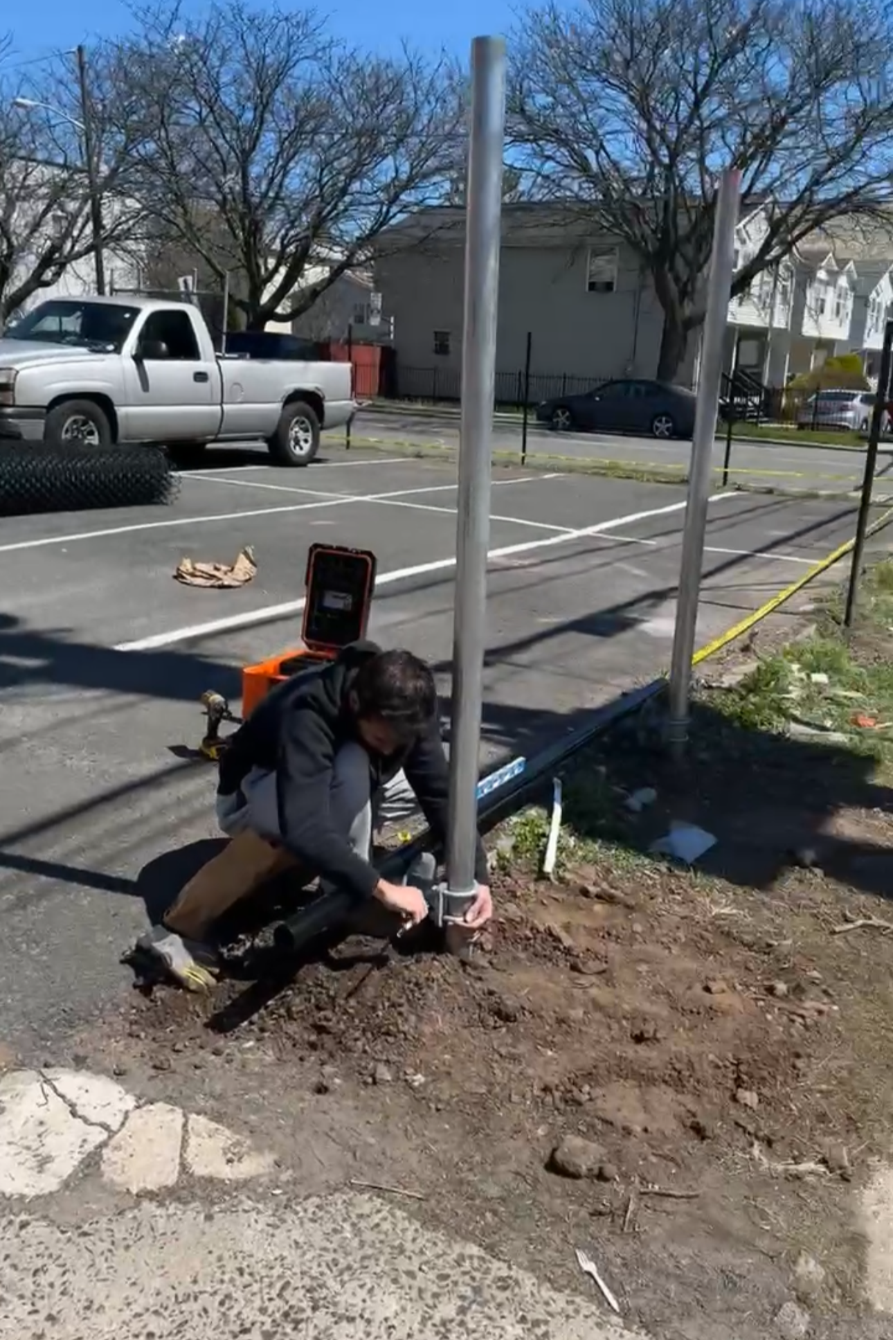 A person installing a pole in the ground at an outdoor parking lot with trees, houses, and a clear blue sky in the background.