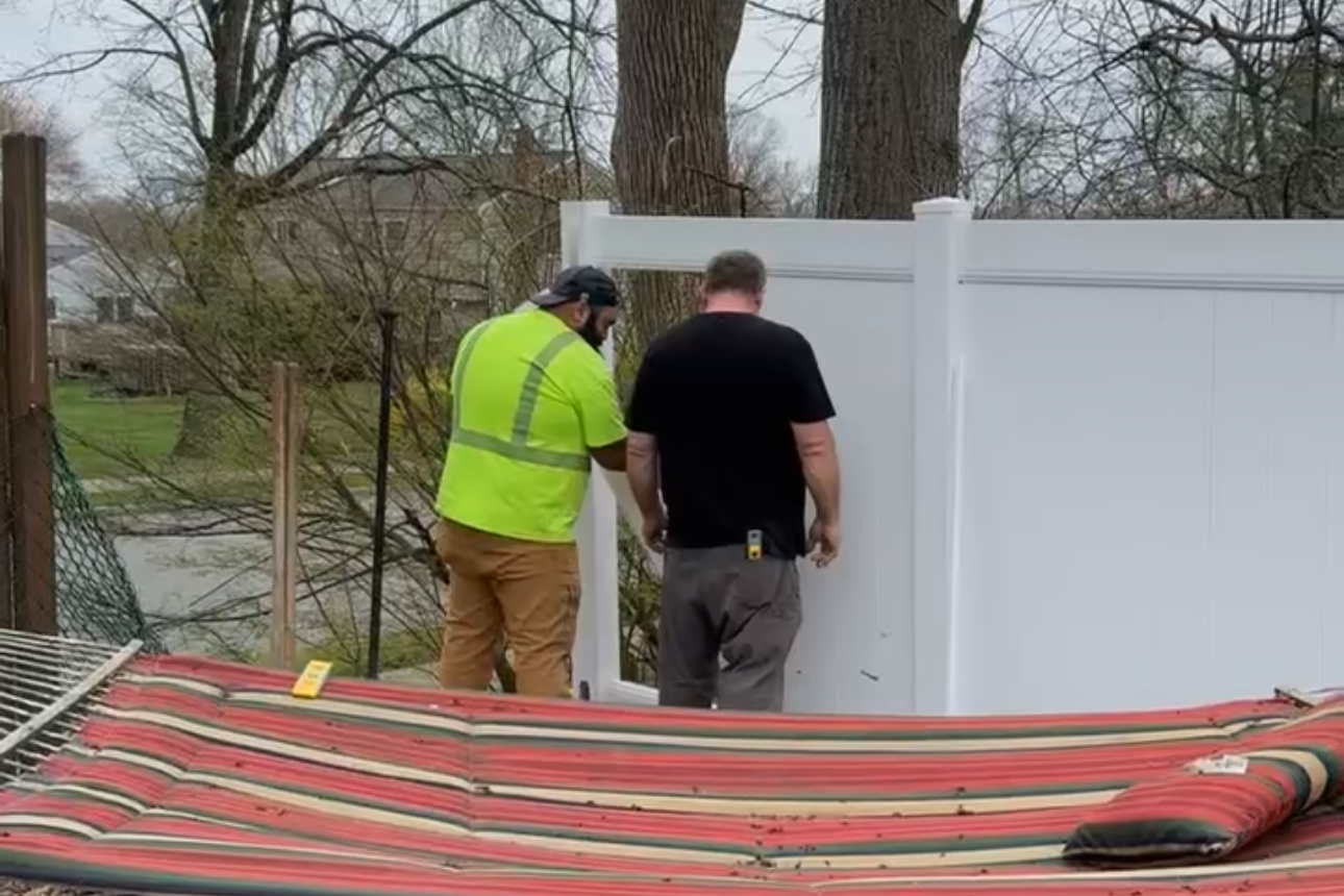 Two men are working to install a white vinyl fence panel in a backyard, with trees and houses in the background.