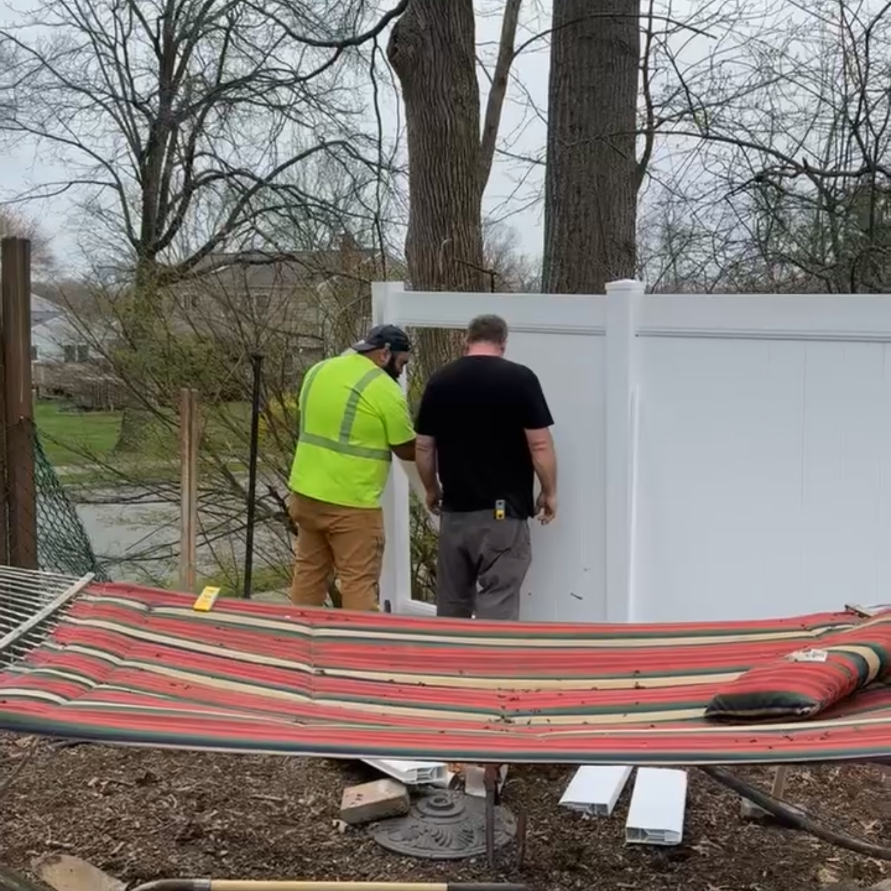 Two men working on installing or repairing a white fence in a backyard, with a large tree and leafless trees in the background, and a striped outdoor mat in the foreground.