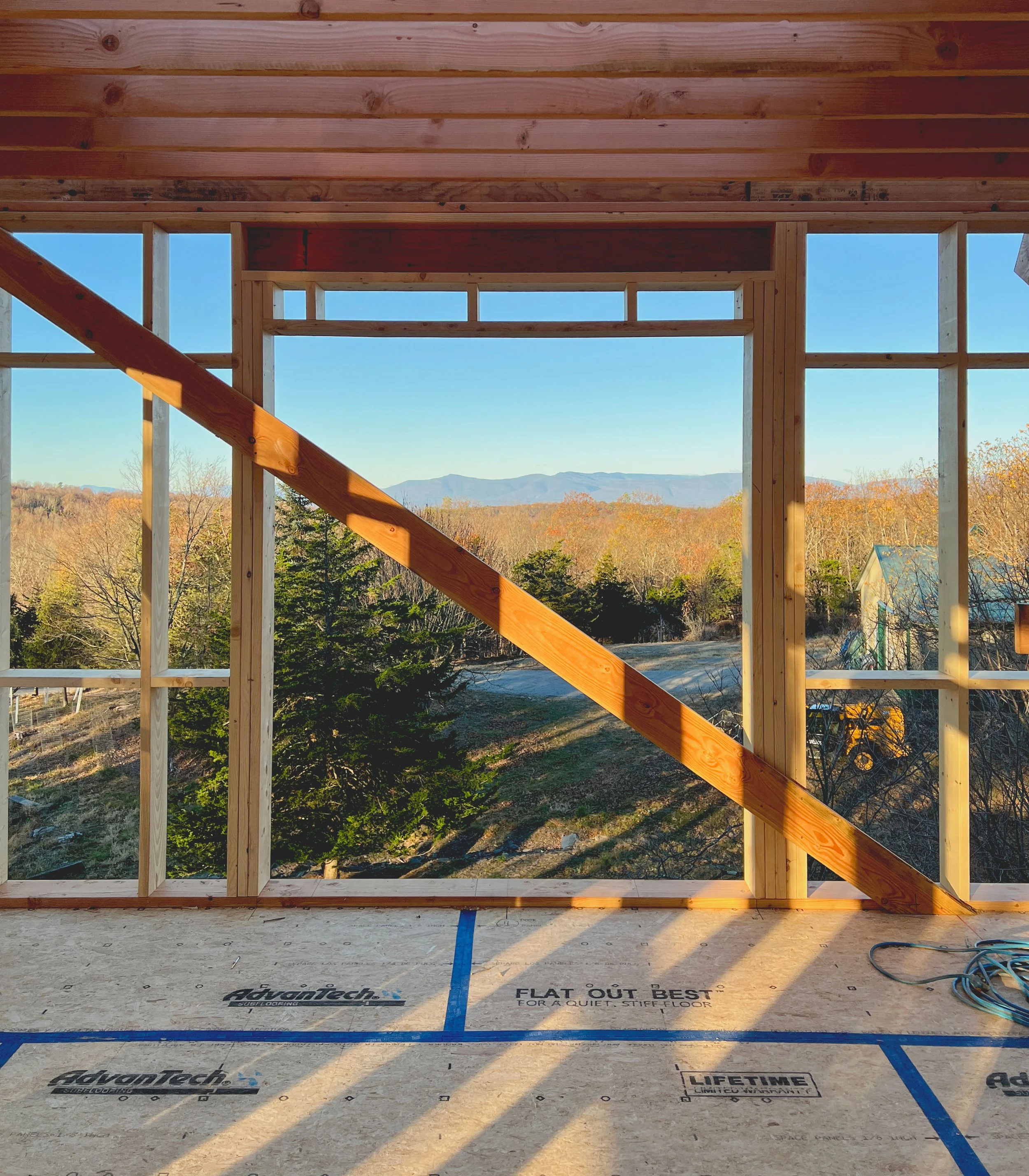 Construction site with a wooden frame of a building, overlooking a scenic landscape with trees and mountains in the distance.