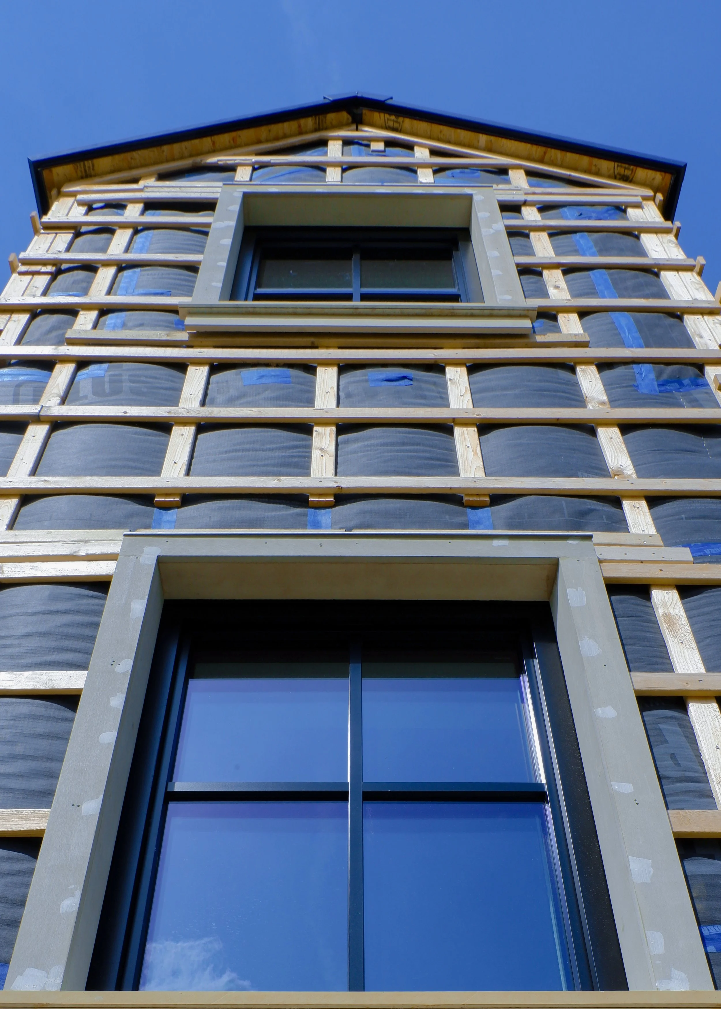 The image shows a building under construction with a window and some insulation wrapped in plastic, with wooden framing and a blue sky in the background.