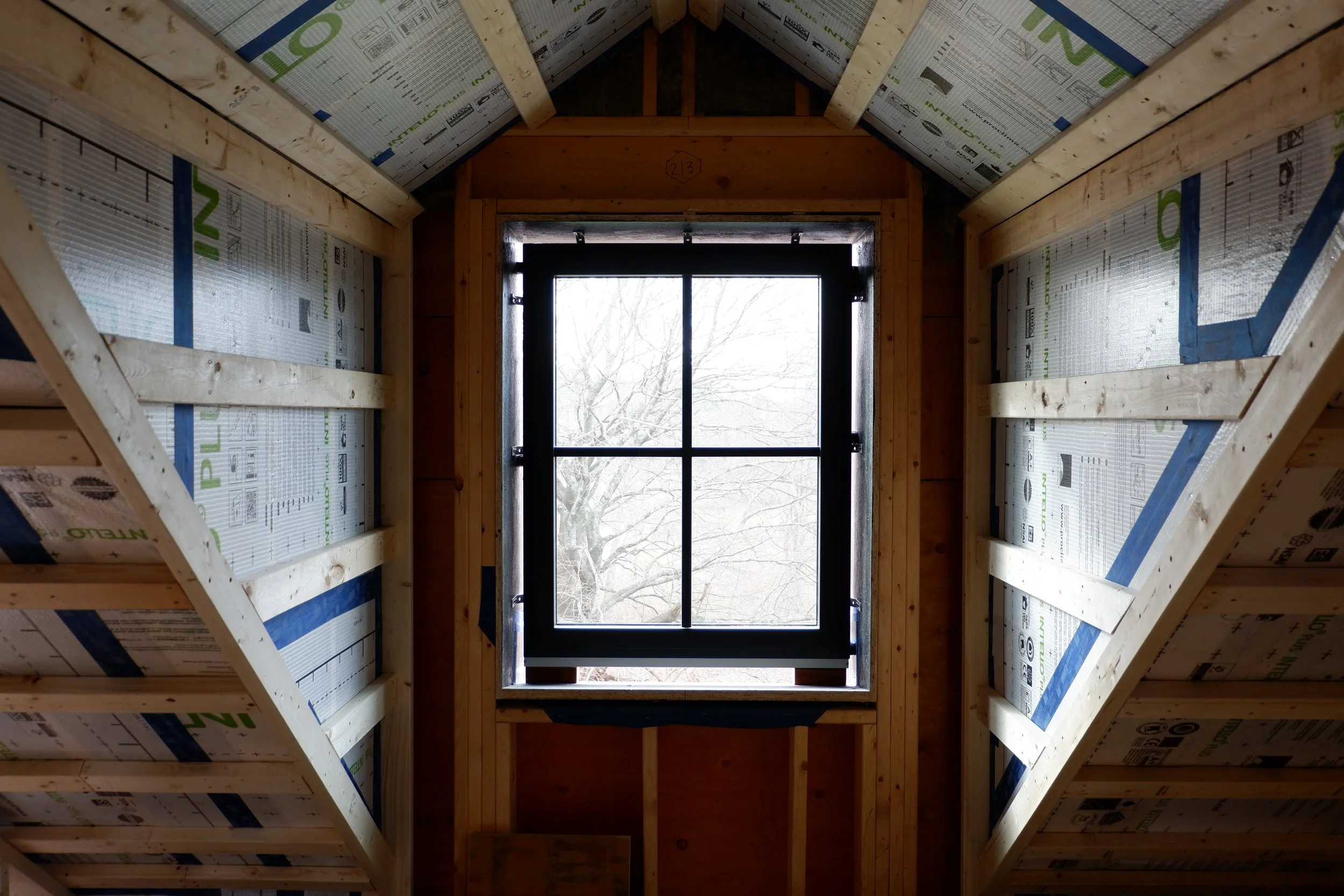 Interior view of a roof under construction with visible wooden beams and insulation material, and a large window at the end showing trees outside.