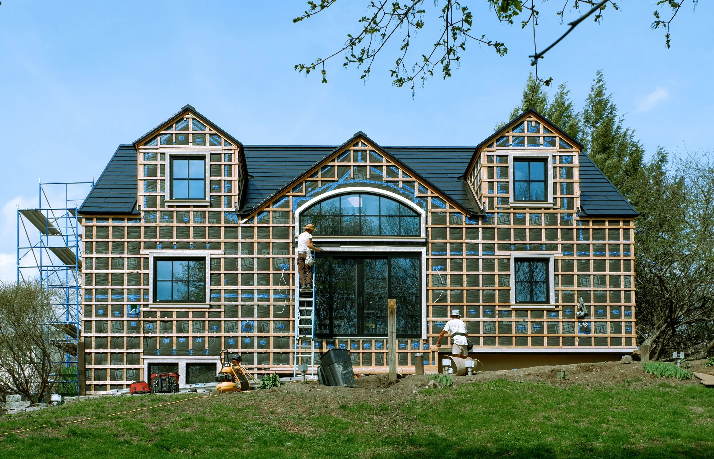 A house under construction with scaffolding, workers installing windows, and a partially finished exterior with black roofing tiles and wood framing visible.