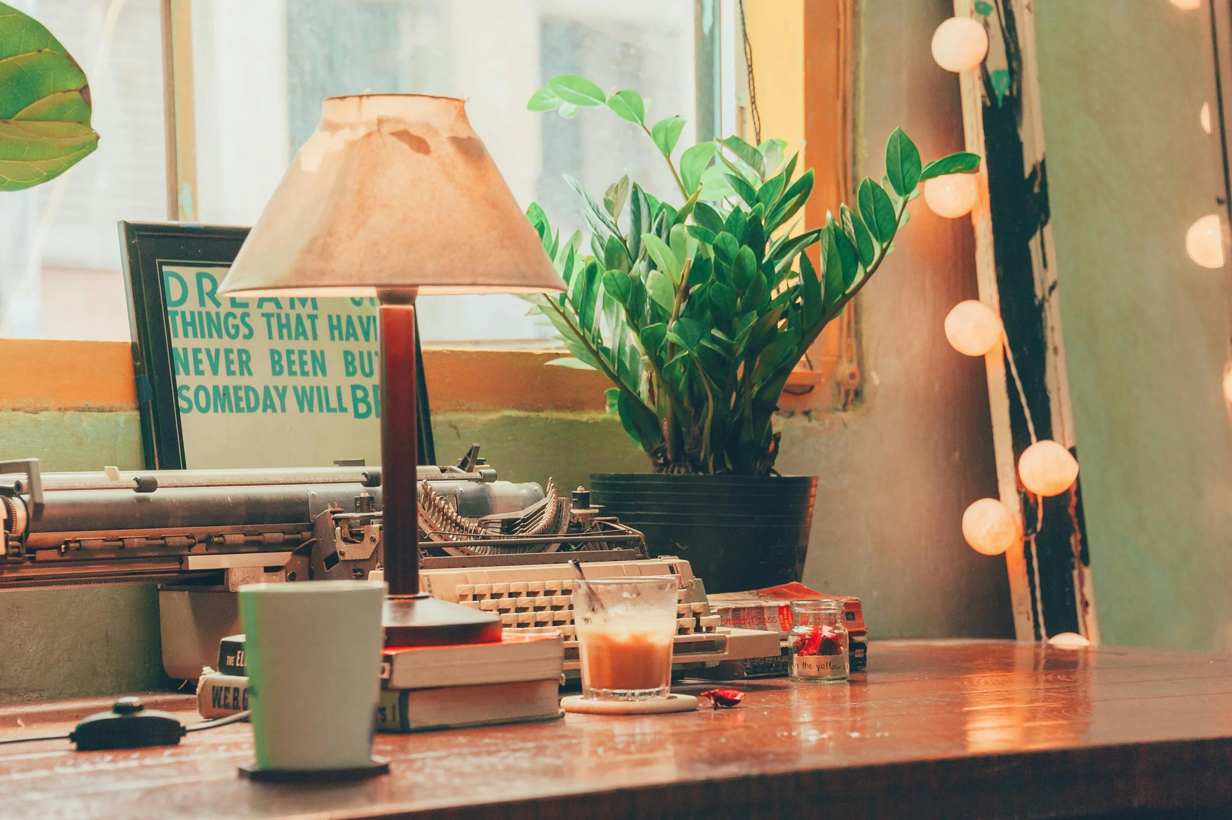 A writer's workstation filled with a thriving plant, a soft lamp, an iced coffee, and a typewriter.