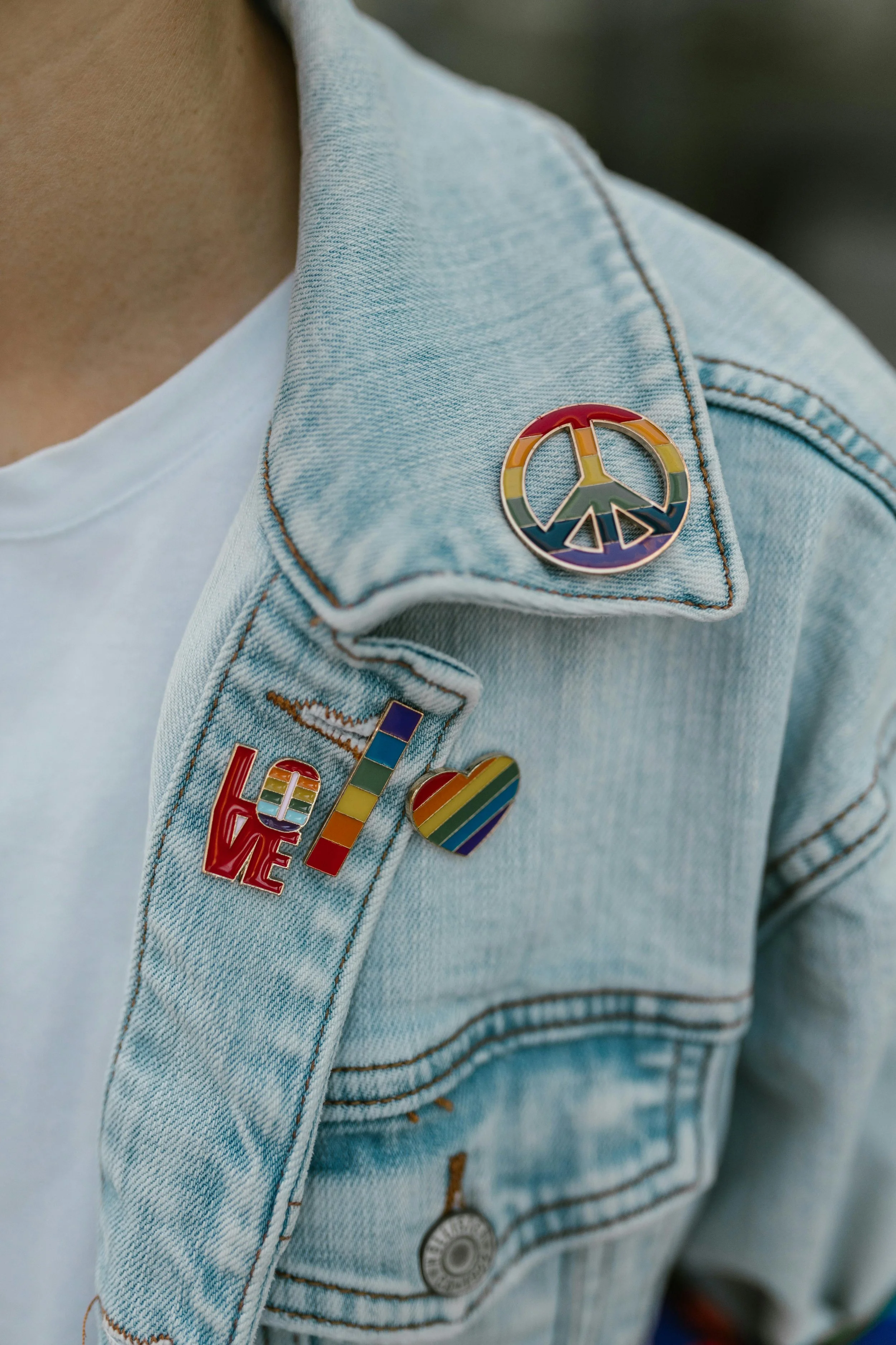 A light blue jean jacket displays multiple rainbow pins promoting peace, equality, and pride.