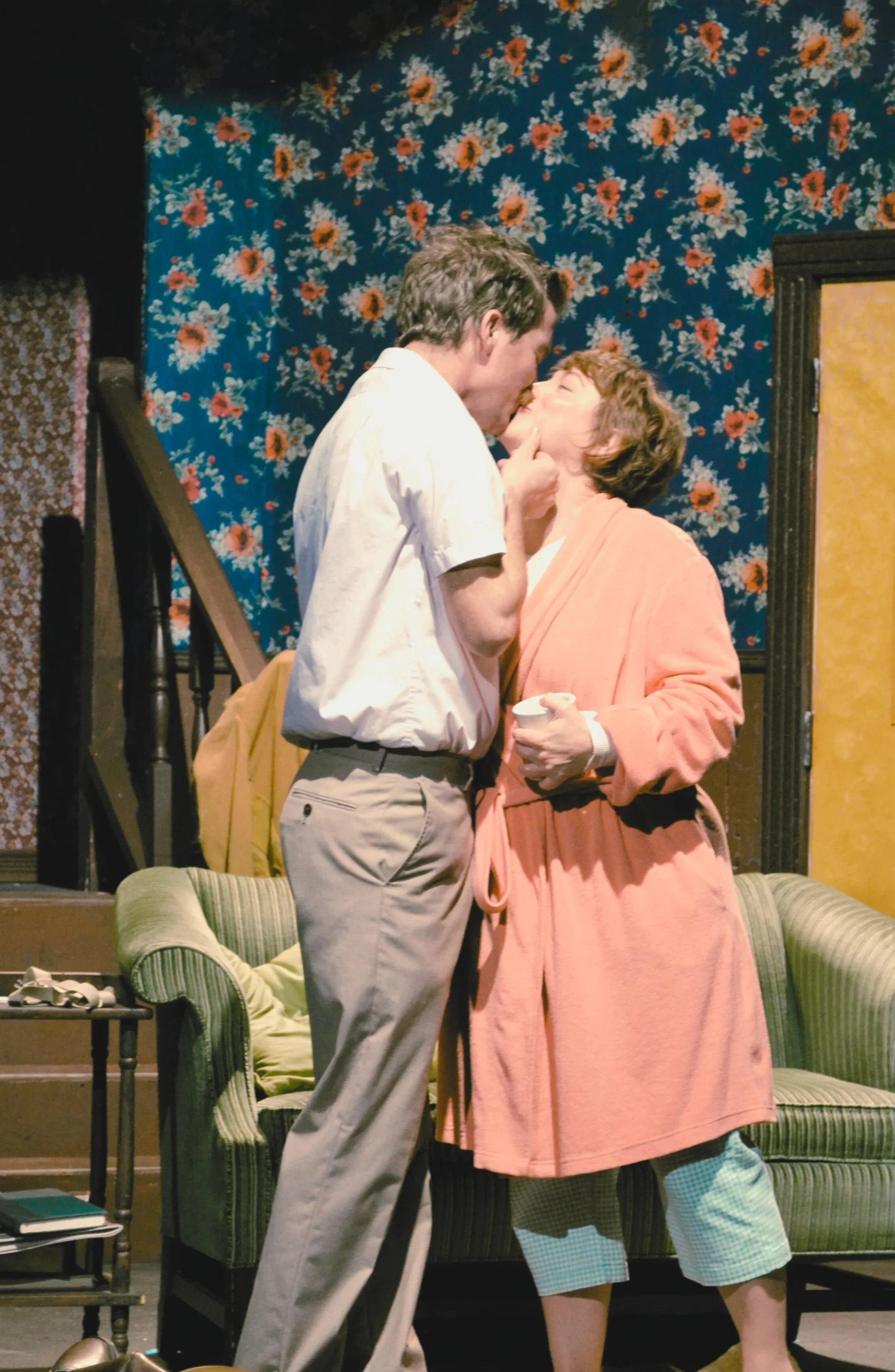 A man in a white shirt gently kisses his wife, in a 1960s theatre set.
