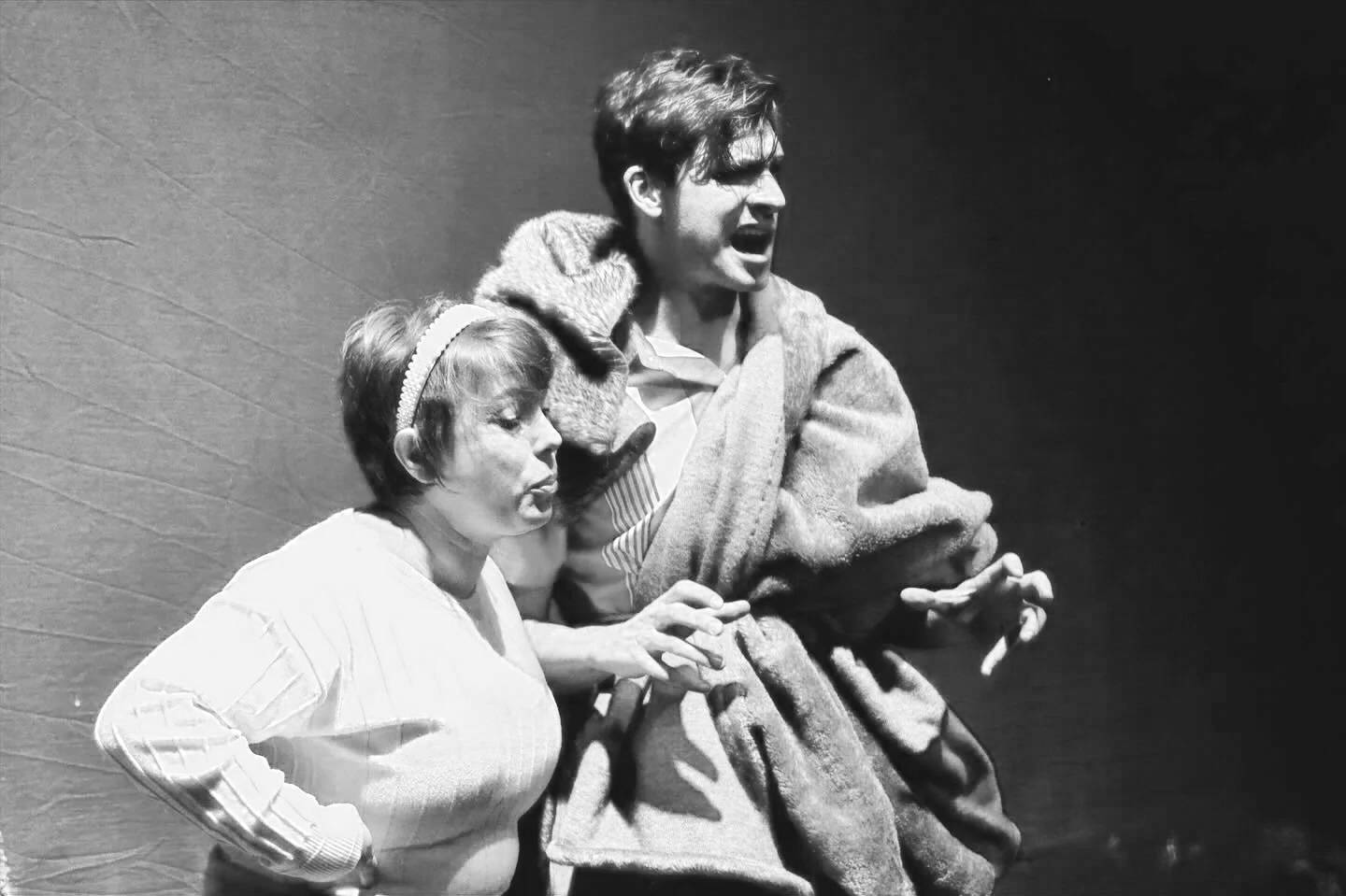 A black and white production still reveals a tall man wrapped in a fur rug and a short woman growling beside him.