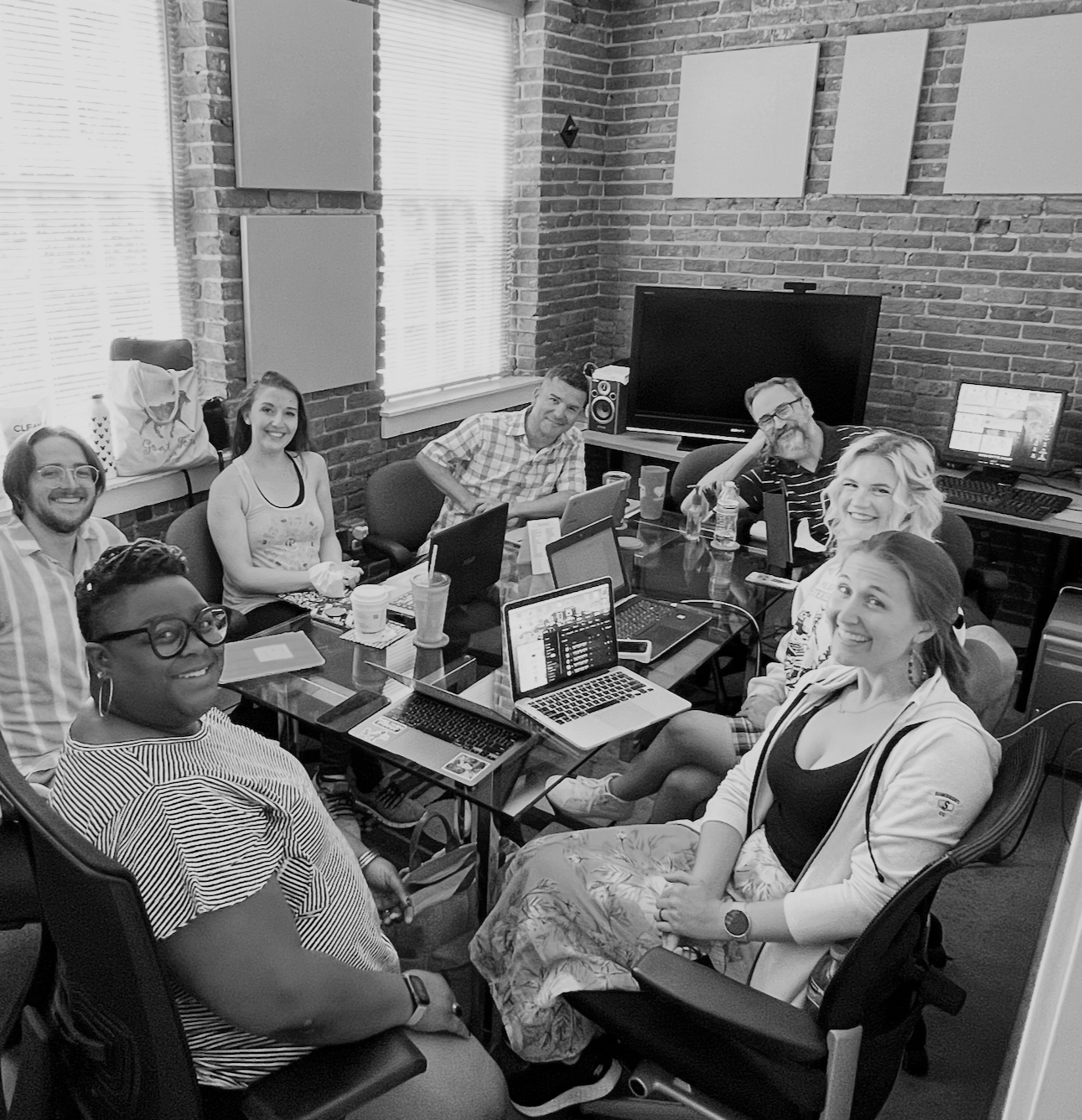A black and white cast photo showing actors around a meeting table, rehearsing.